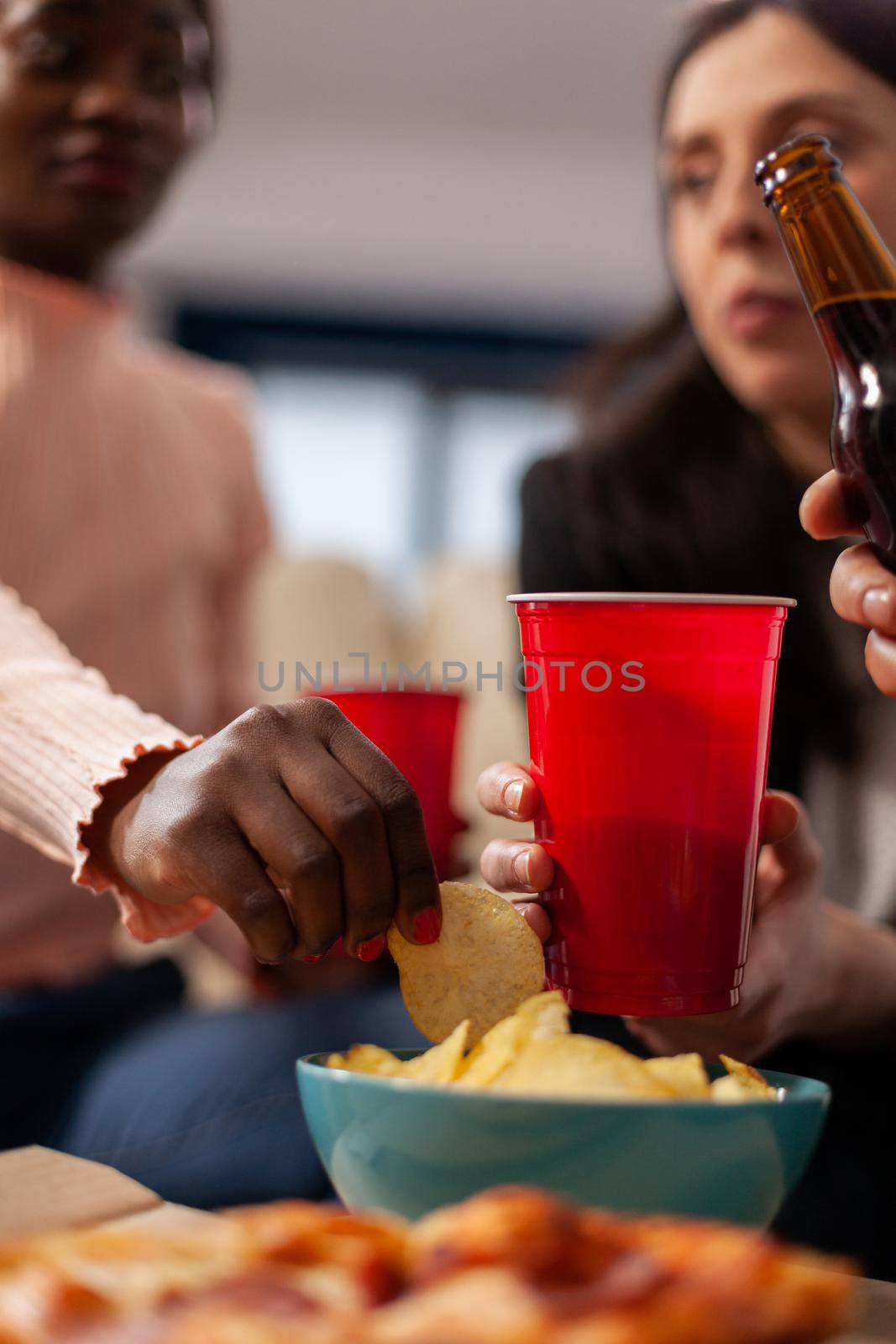 Close up of cups of beer and chips on table at office after work while multi ethnic friends celebrate at indoors party. Diverse group of workers have fun with snacks pizza bottles of alcohol