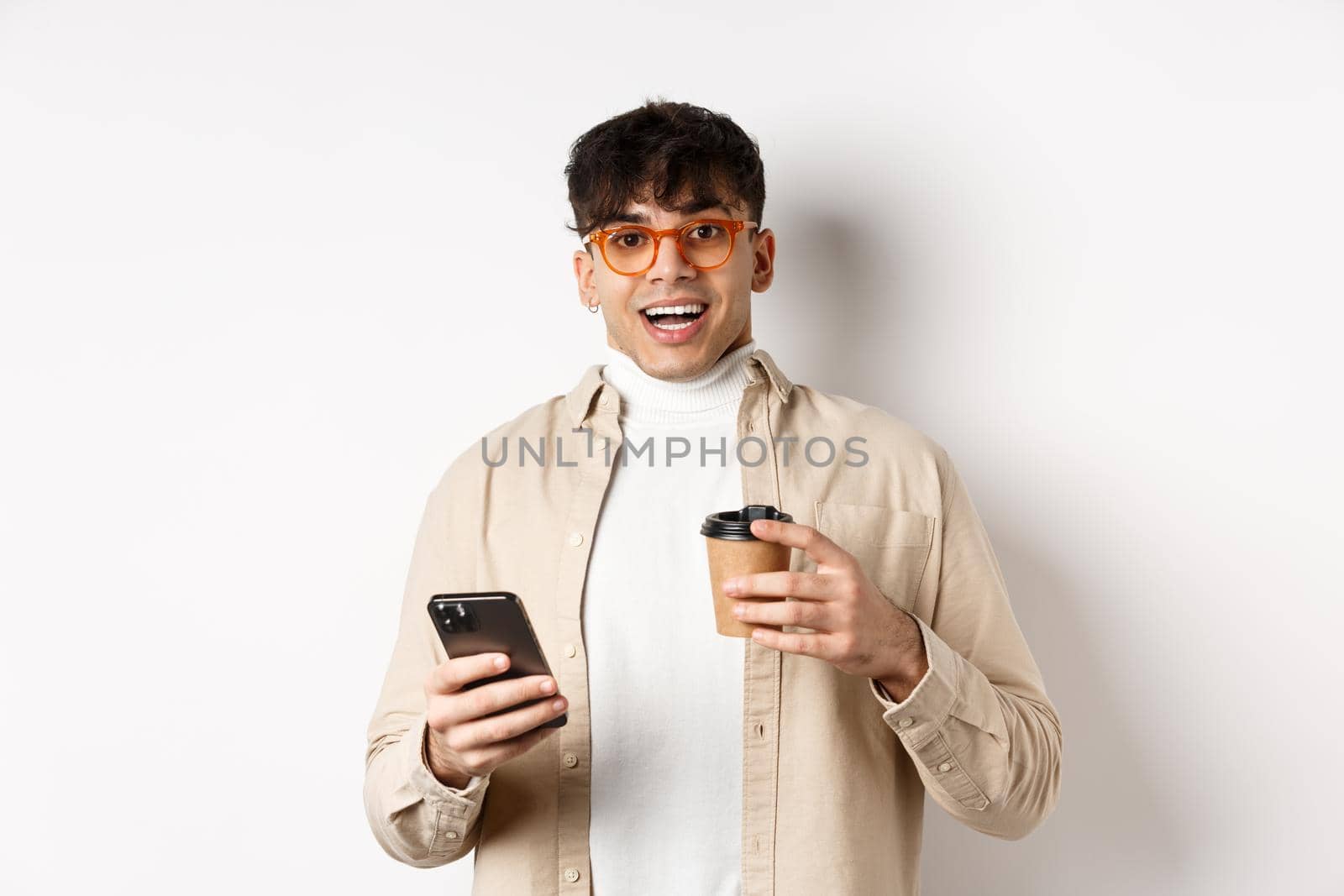 Cheerful smiling young man in glasses drinking coffee and holding smartphone, looking excited at camera, standing on white background by Benzoix