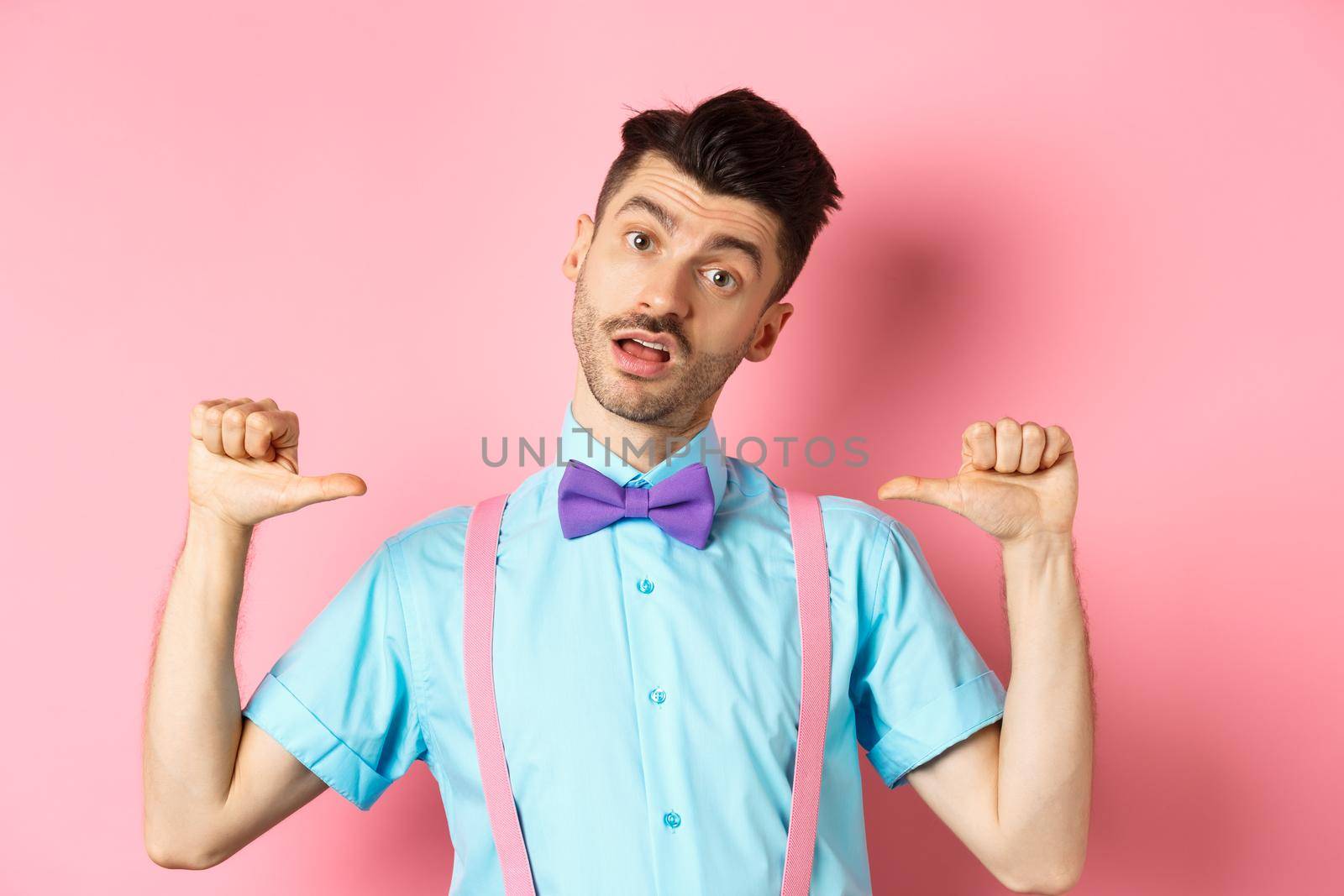 Sassy and confident young man in suspenders and bow-tie, pointing at himself and looking like professional, standing over pink background.