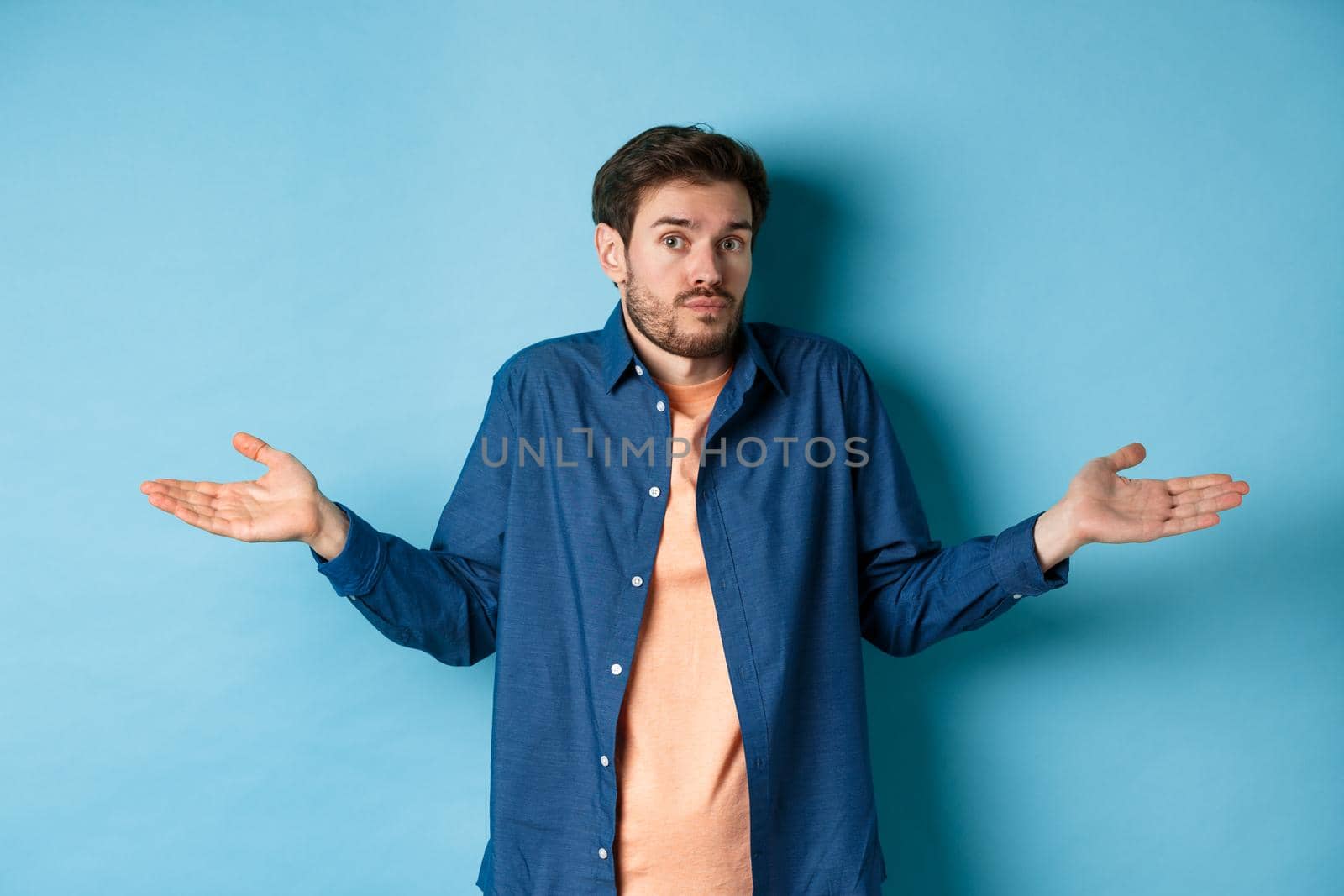 Confused young man with beard, shrugging shoulders and looking clueless at camera, know nothing, standing on blue background.