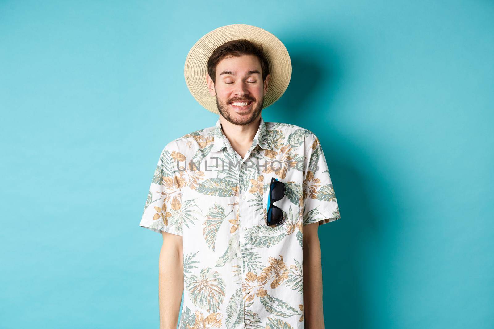 Happy tourist enjoying summer holiday, wearing straw hat and hawaiian shirt, standing joyful on blue background.