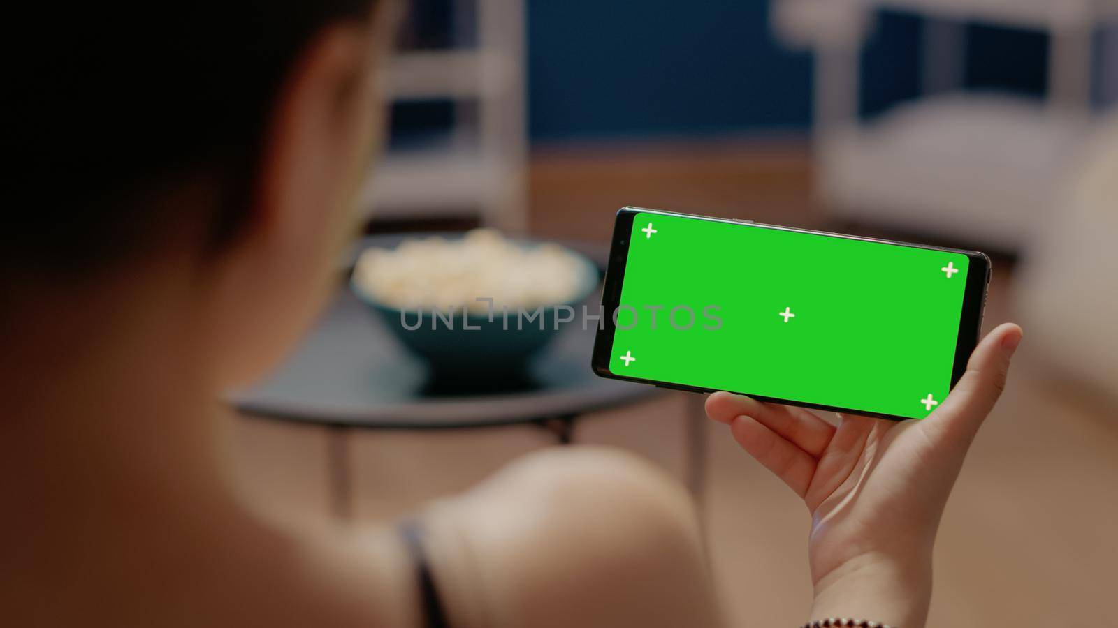 Close up of mobile phone with horizontal green screen on display. Young woman sitting at home looking at blank isolated background and mockup virtual template for chroma key and copy space