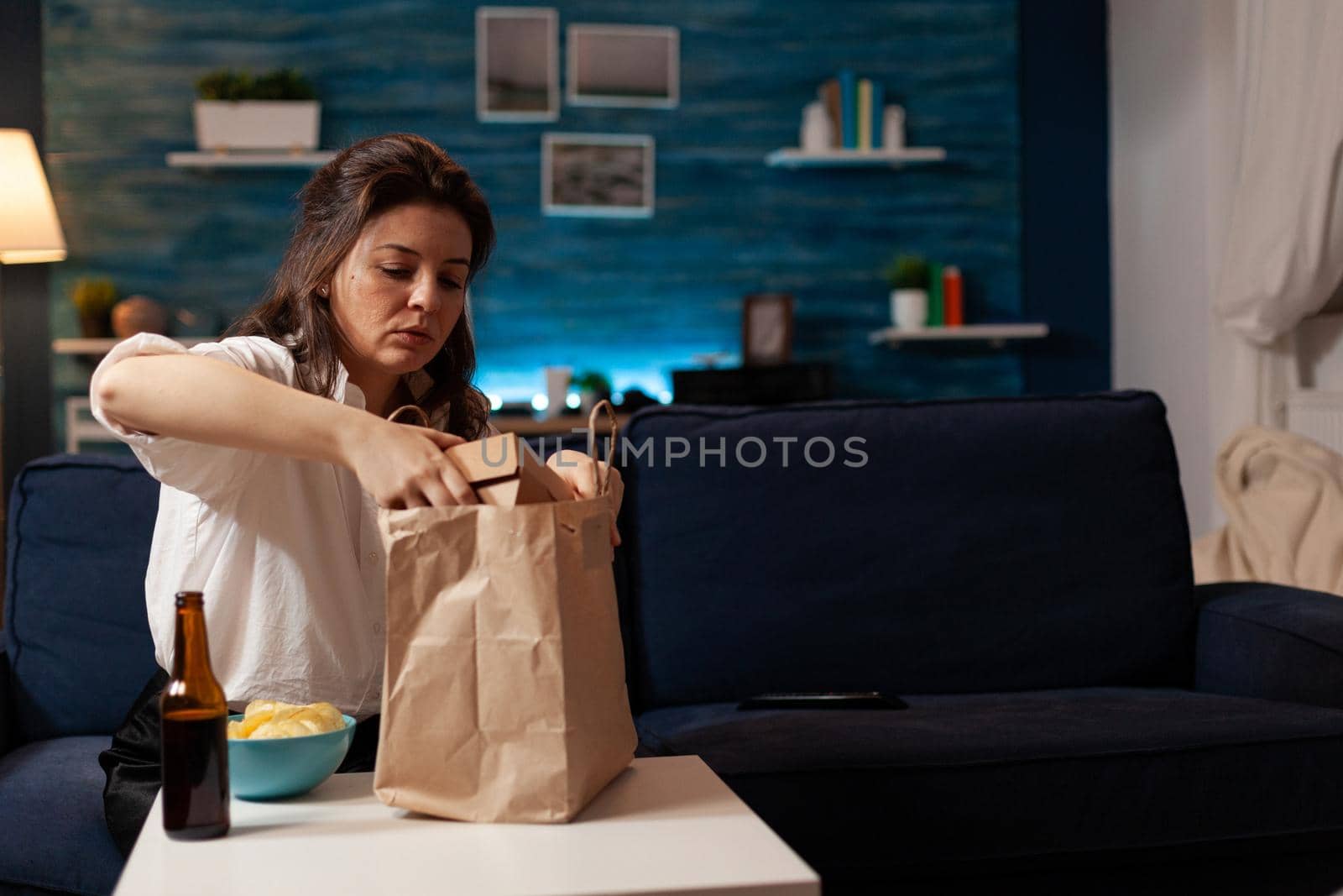 Smiling cheerful woman unpacking fast food meal delivery sitting on sofa during junk-food home-delivered in living room. Caucaisan female enjoying takeaway service lunch in evening