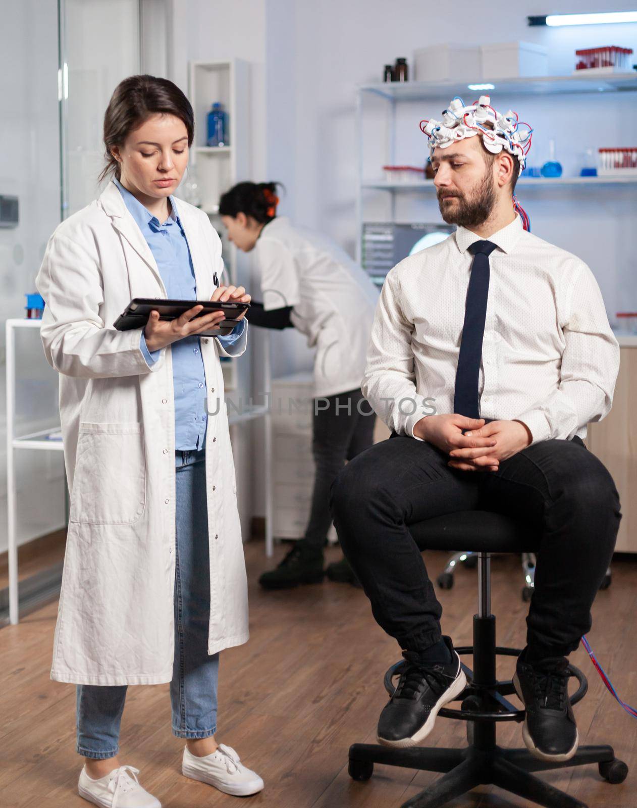 Specialist doctor in neuroscience taking notes on clipboard while testing brain functions of man using eeg headset, treating nervous system dysfunctions in moden laboratory.