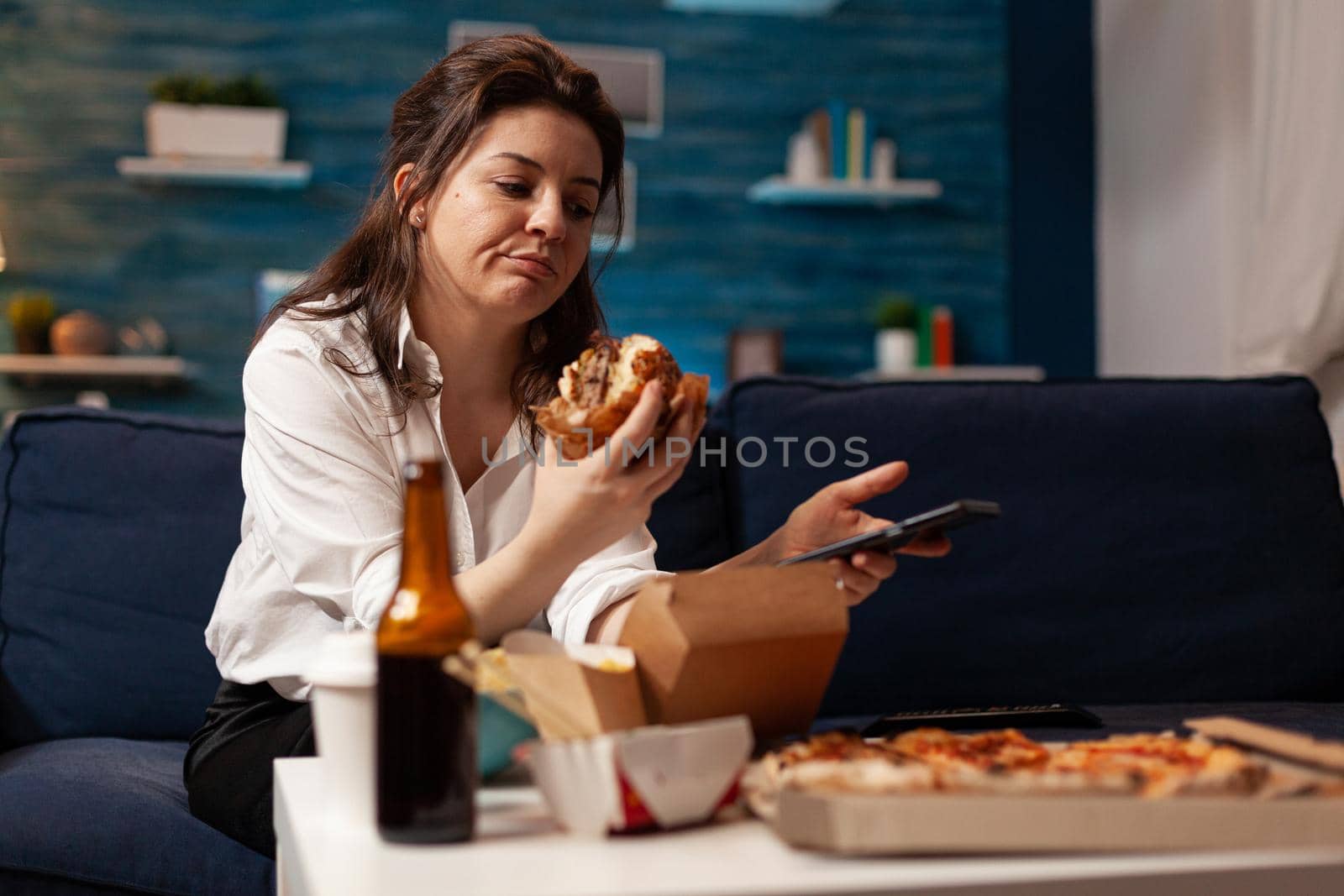 Caucasian female holding tastu burgery in hands browsing on social media using smartphone relaxing on couch during junck-food night. Woman enjoying takeaway food home delivery