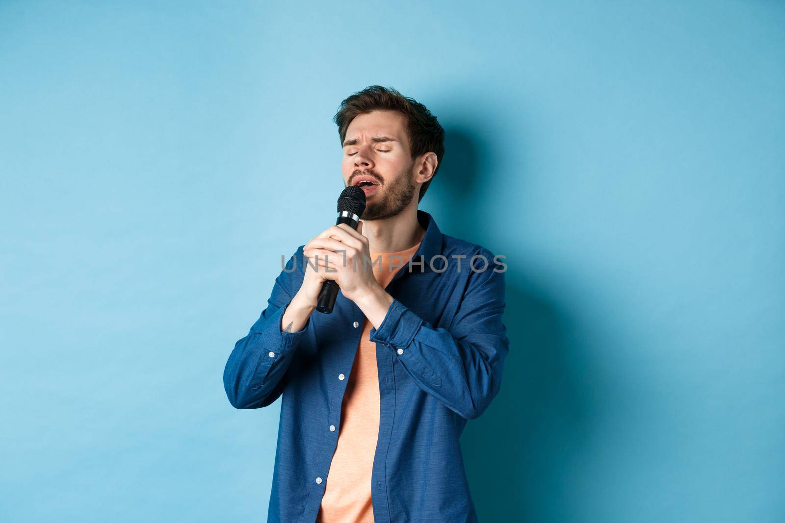 Romantic guy singing in microphone, performing on stage, standing on blue background.