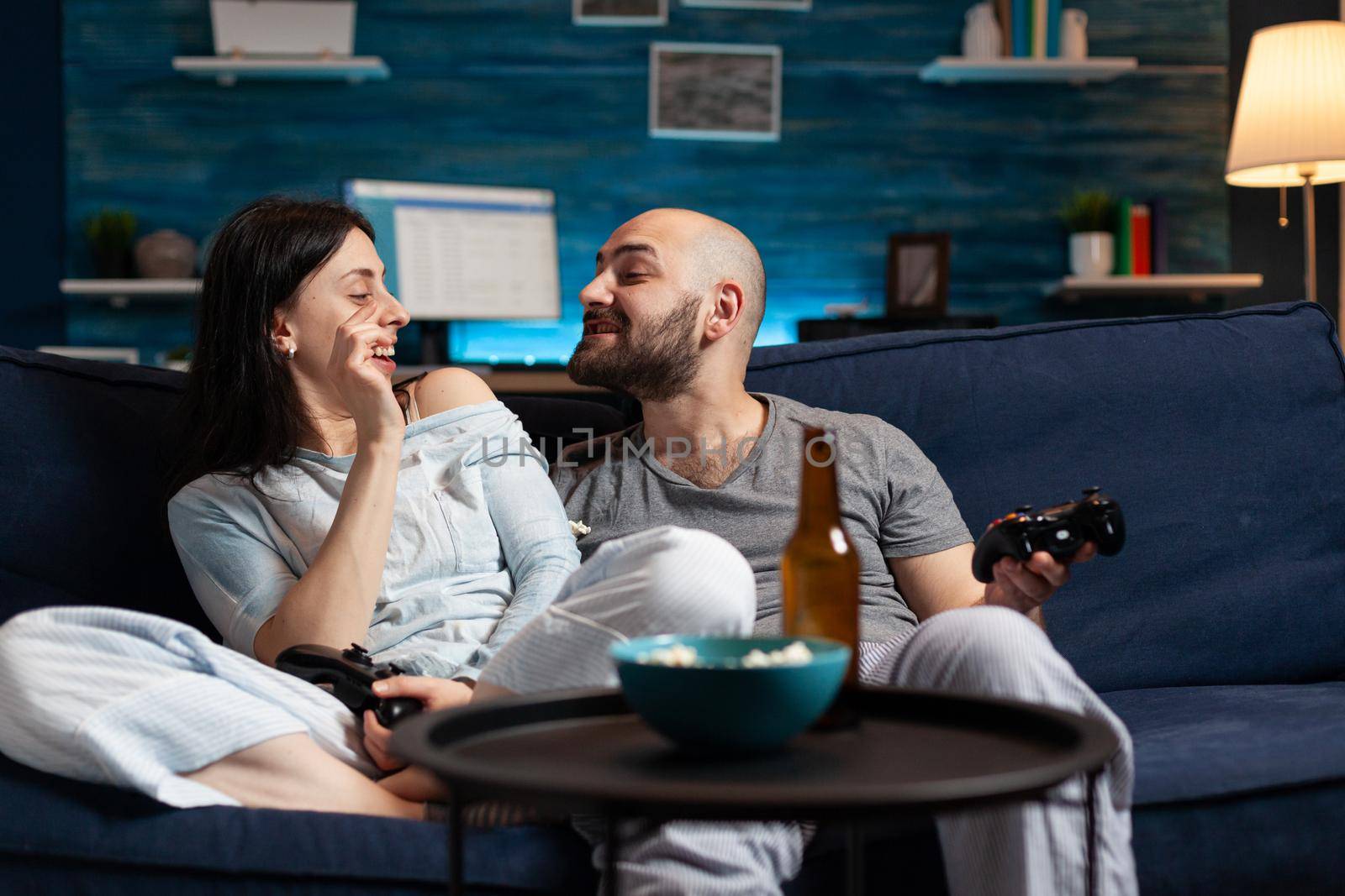 Funny relaxed couple sitting on couch relaxing at night laughing having fun eating popcorn. Happy people enjoying time playing online video games using joystick, happiness and married people concept.