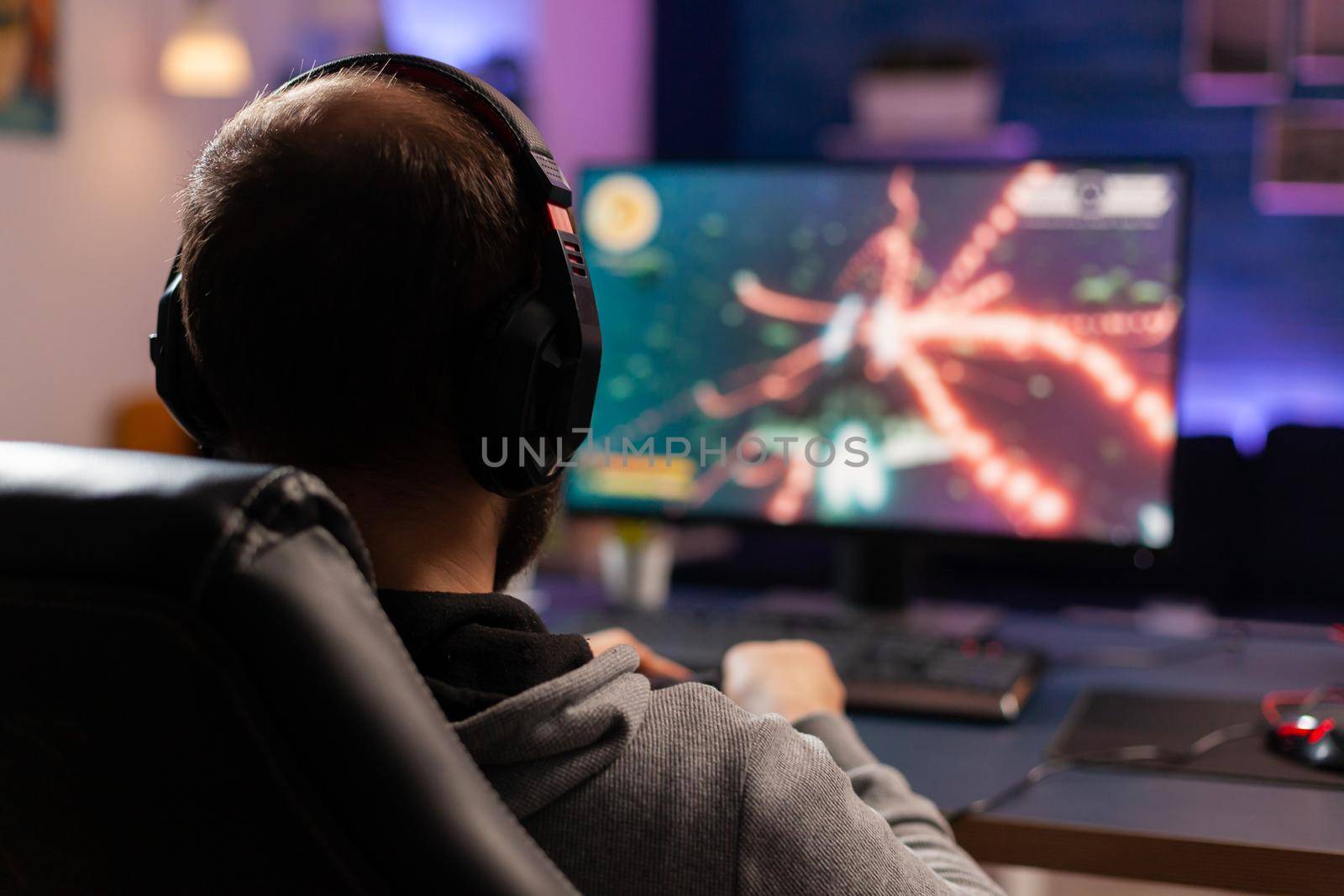 Back view shot of concentrate gamer streaming online videogames on computer using wireless controller. Player man with headphones playing games in room with neon light and professional equipment
