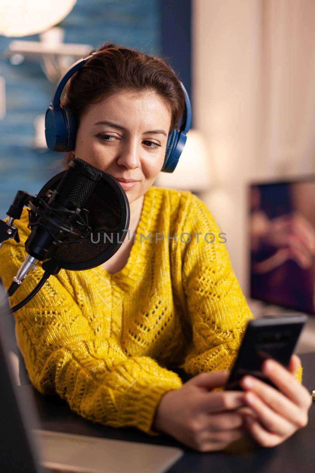 Happy blogger reading messages of fans using smartphone sitting in home podcast studio during livestreaming, recording. Online show On-air production internet broadcast host streaming live content.