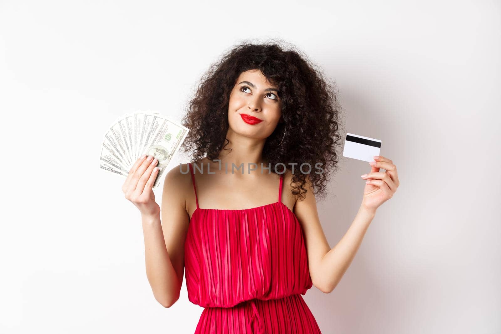 Shopping. Woman thinking and smiling, holding money with plastic credit card, wearing red elegant dress and evening makeup, white background.