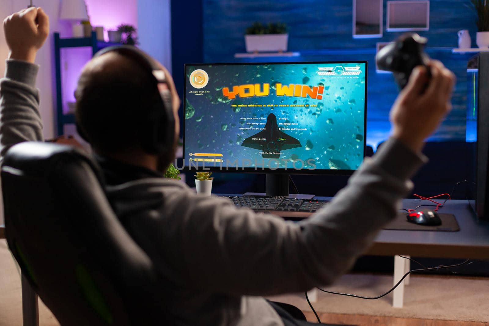 Addicted man playing space shooter videogame using headphones and wireless console. Excited gamer raising hands after winning online championship game at home gaming studio