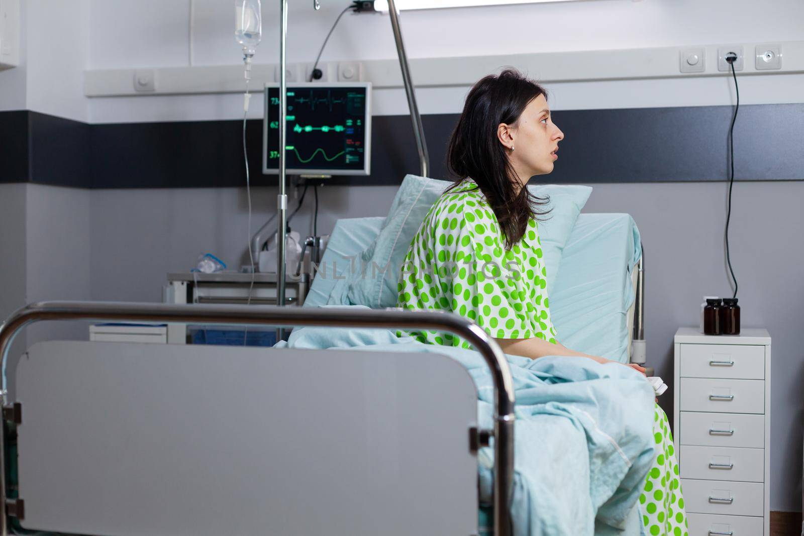 Hospitalized sick woman with illness recovering after medical surgery sitting on bed in hospital ward during recovery waiting for consultation. Patient healthcare treatment in modern clinic