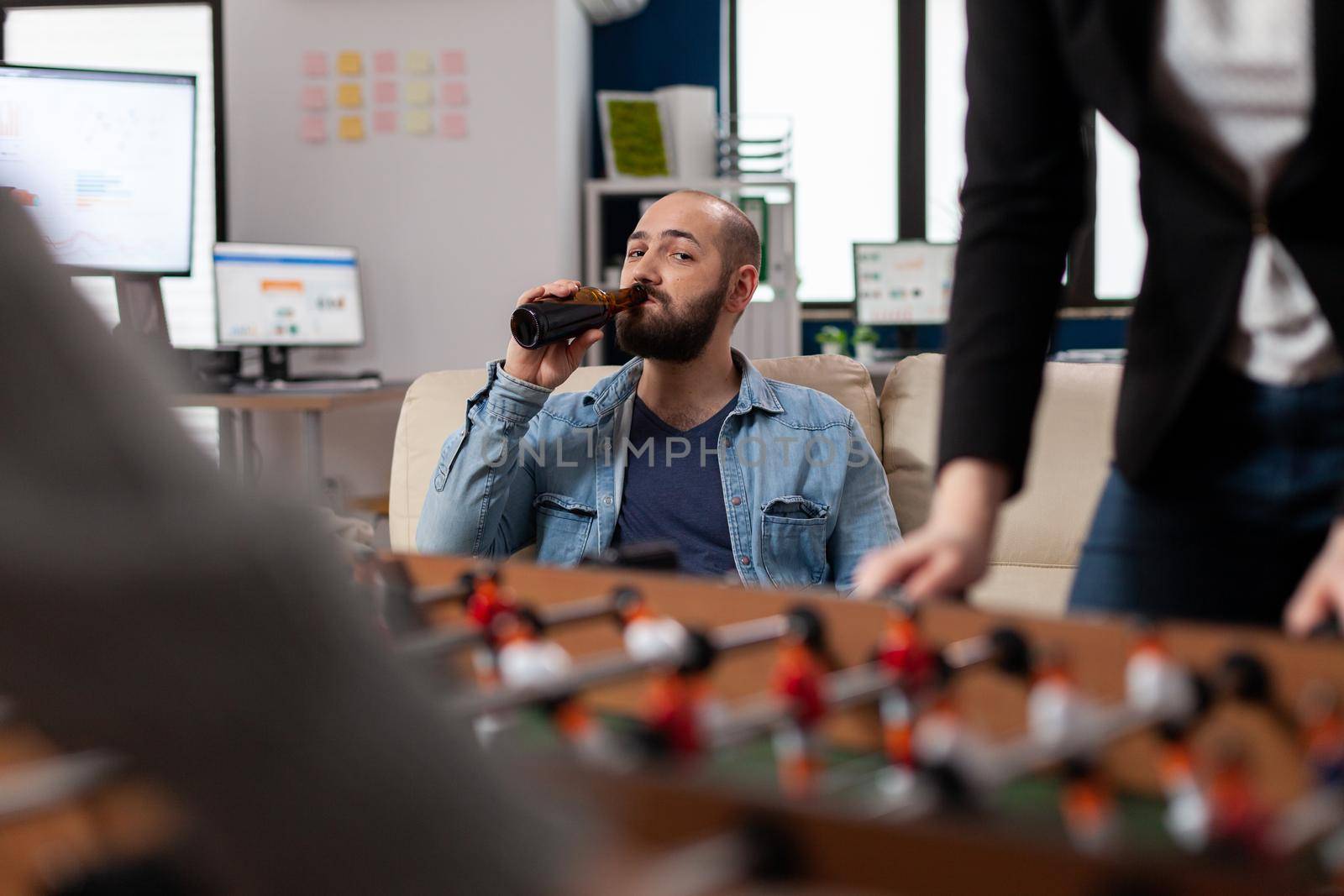 After work man drinking alcohol at foosball table soccer football watching toy game. Caucasian business worker play at office for enjoyment holding bottle of beer having fun entertainment
