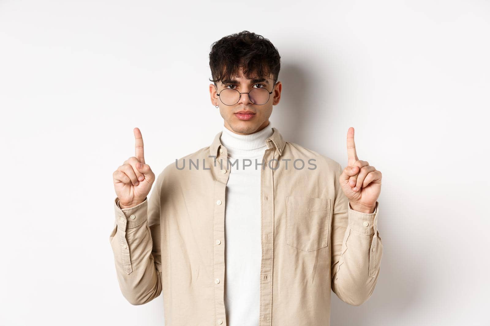 Handsome hipster guy in glasses pointing fingers up at empty space, looking serious at camera, standing on white background.