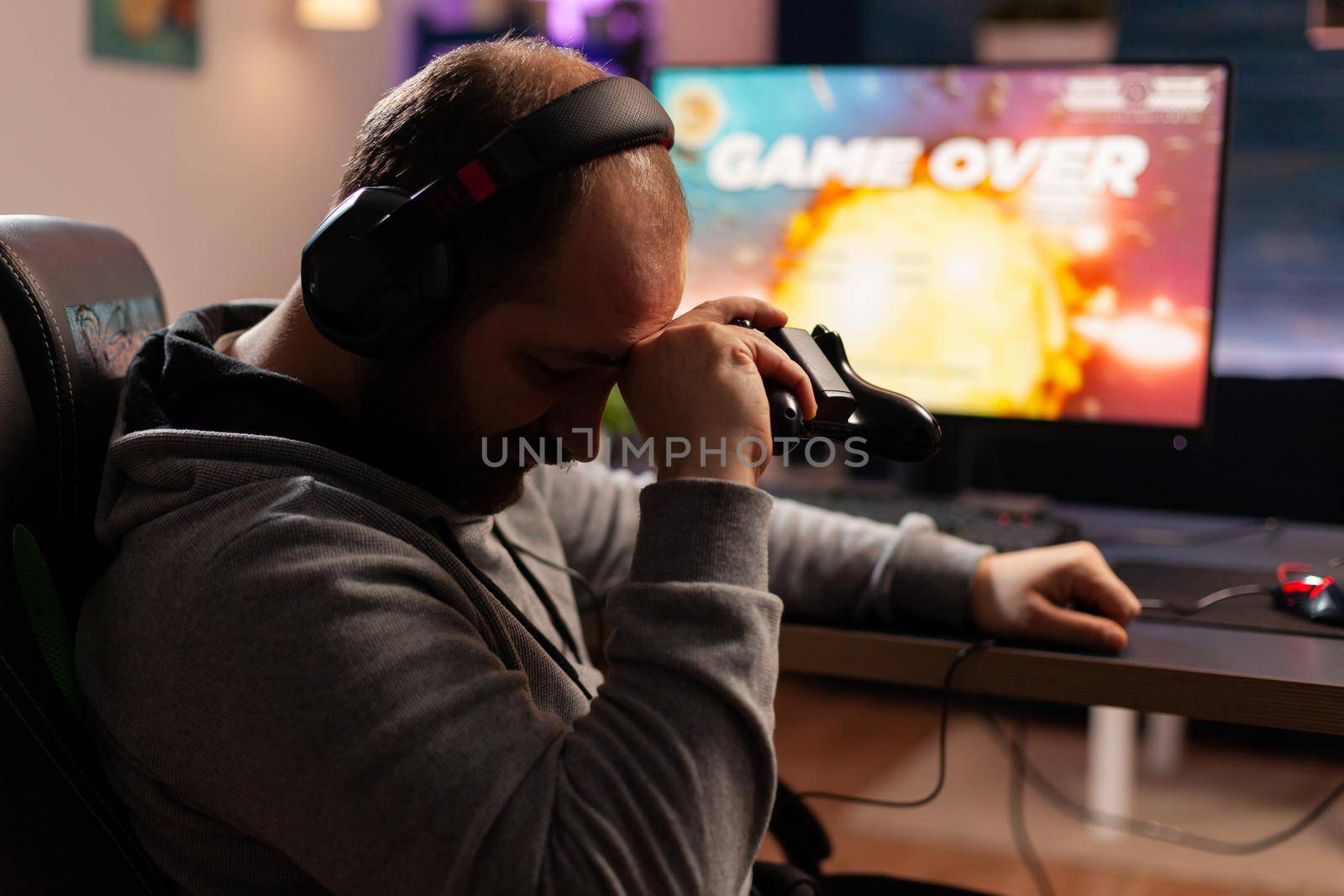 Upset cyber player losing video game sitting on gaming desk late at night in living room. Man streaming games for online championship using headphones and professional joystick