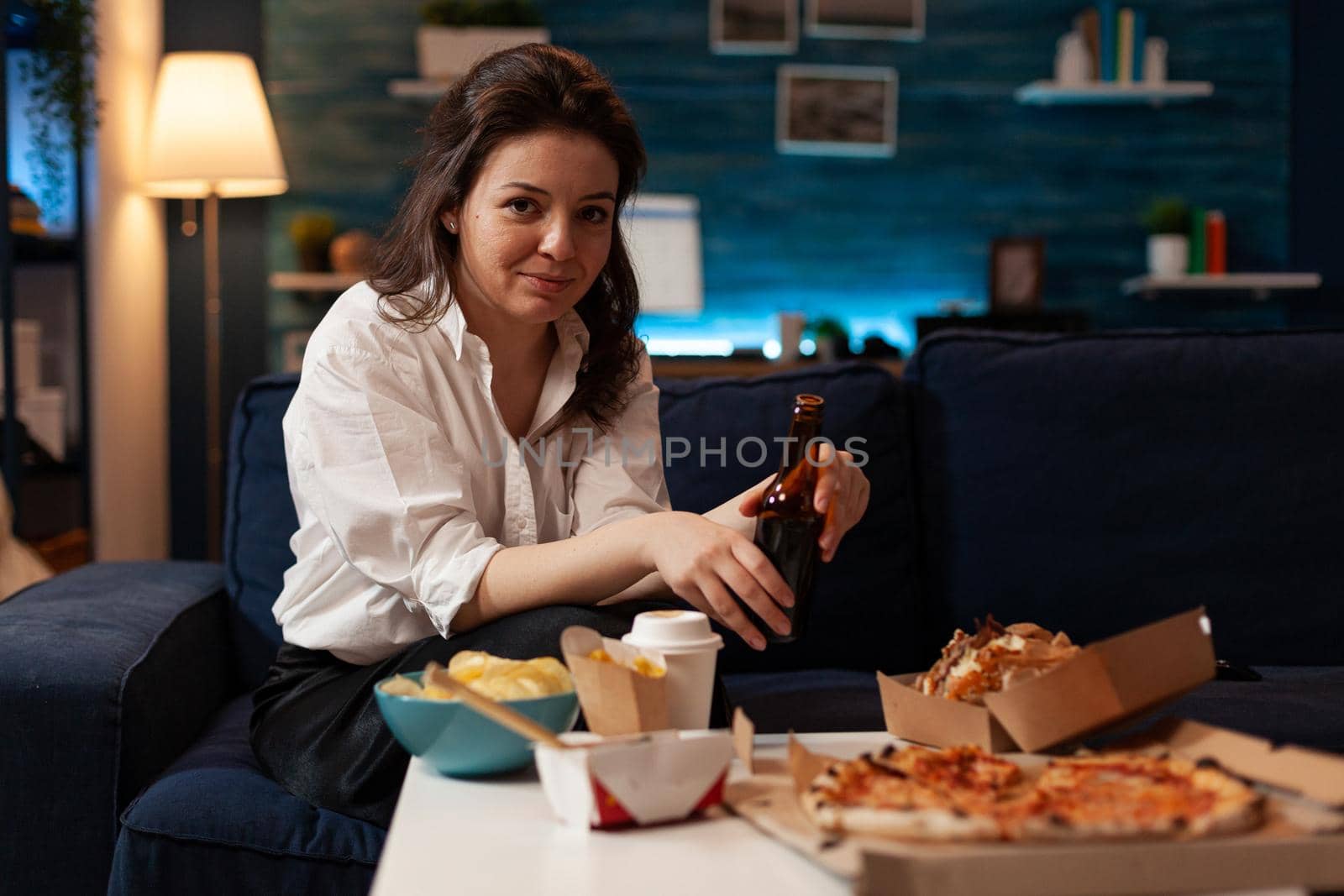 Portrait of cheerful woman sitting on couch smiling at camera while enjoying home-delivered late at night in living room. Caucasian female holding beer bottle. Takeaway food delivery