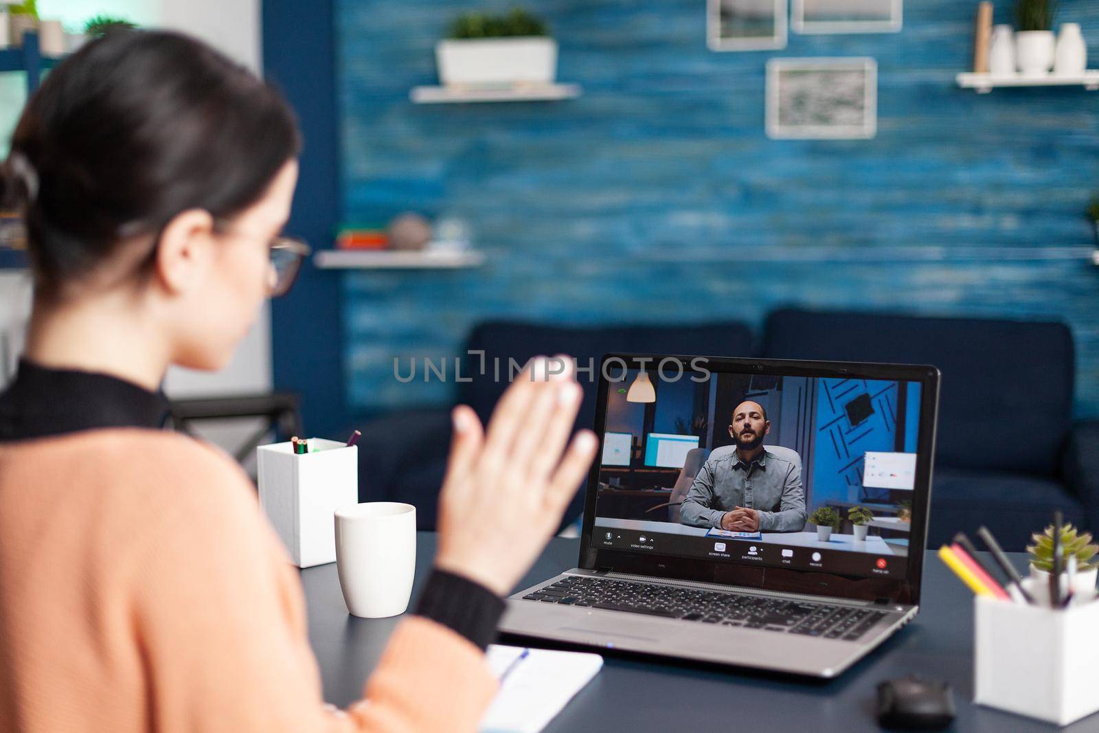 Student waving hand to her university teacher during videocall meeting. Caucasian female discussing about remote education because of coronavirus quarantine while sitting at desk in living room