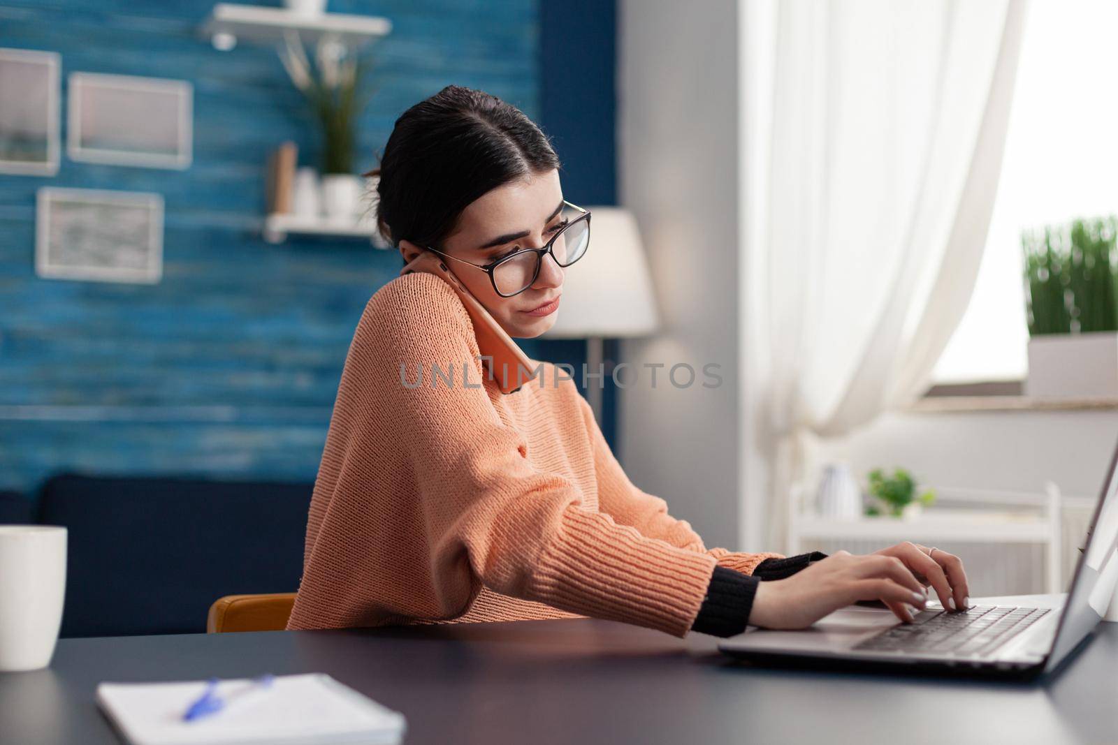 Teenager woman working at e-learning platform for university project during remote education. Student talking at phone about digital courses while sitting at desk in living room