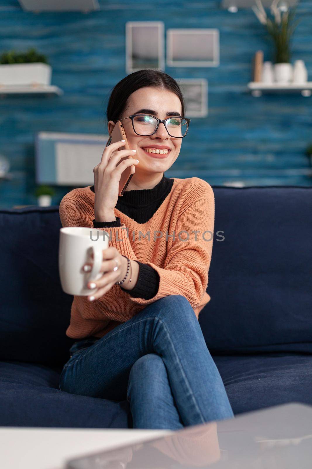 Portrait of teenager talking about lifestyle on modern smartphone while laughing with her friend sitting on couch in living room. Young woman having fun during funny lifestyle conversation call