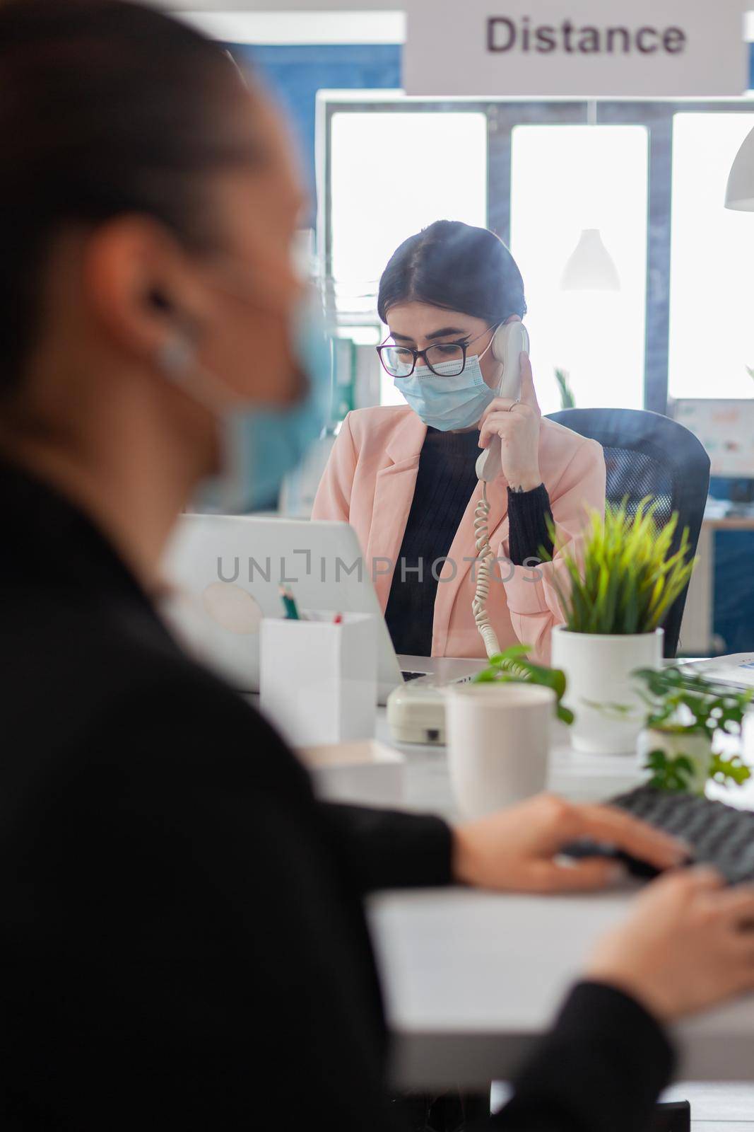 Woman entrepreneur discussing on phone with client, wearing face mask as safety prevention, keeping social distancing with colleague in new normal office during coronavirus flu outbreak.