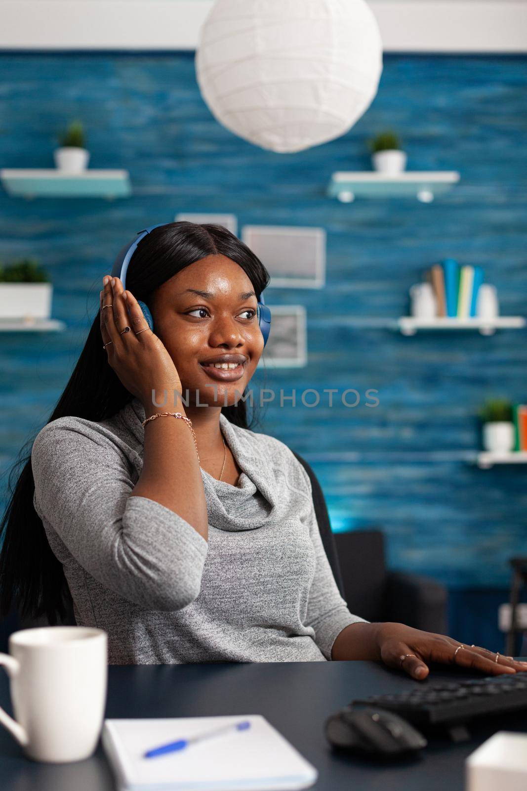 African american student wearing headphone listening online learning course using university platform sitting at desk in living room. Black woman working remote from home during high school class