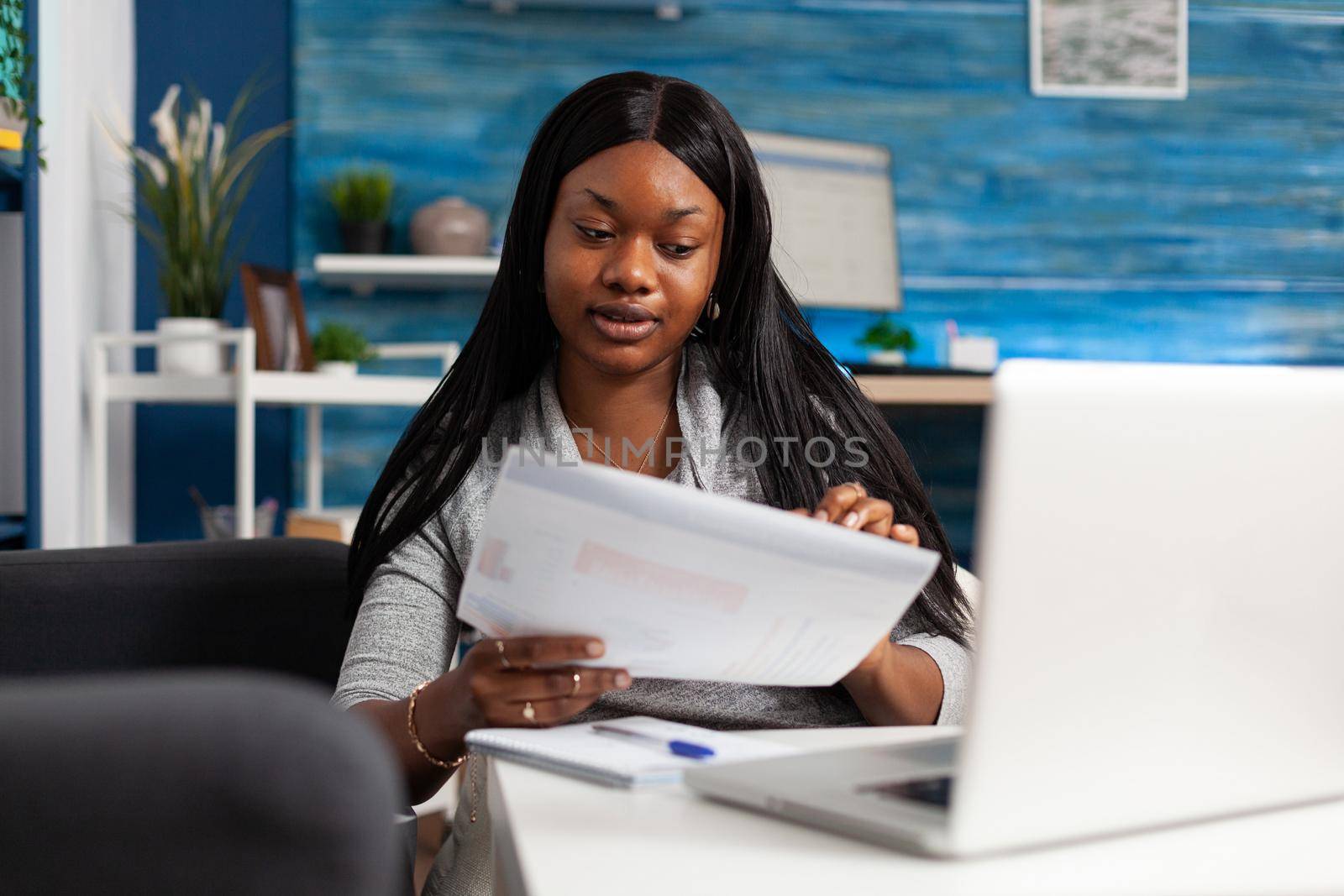 Smart afro american student analyzing accounting infographics planning financial strategy sitting at table in living room. Black woman remote working at marketing statistics checking graph document