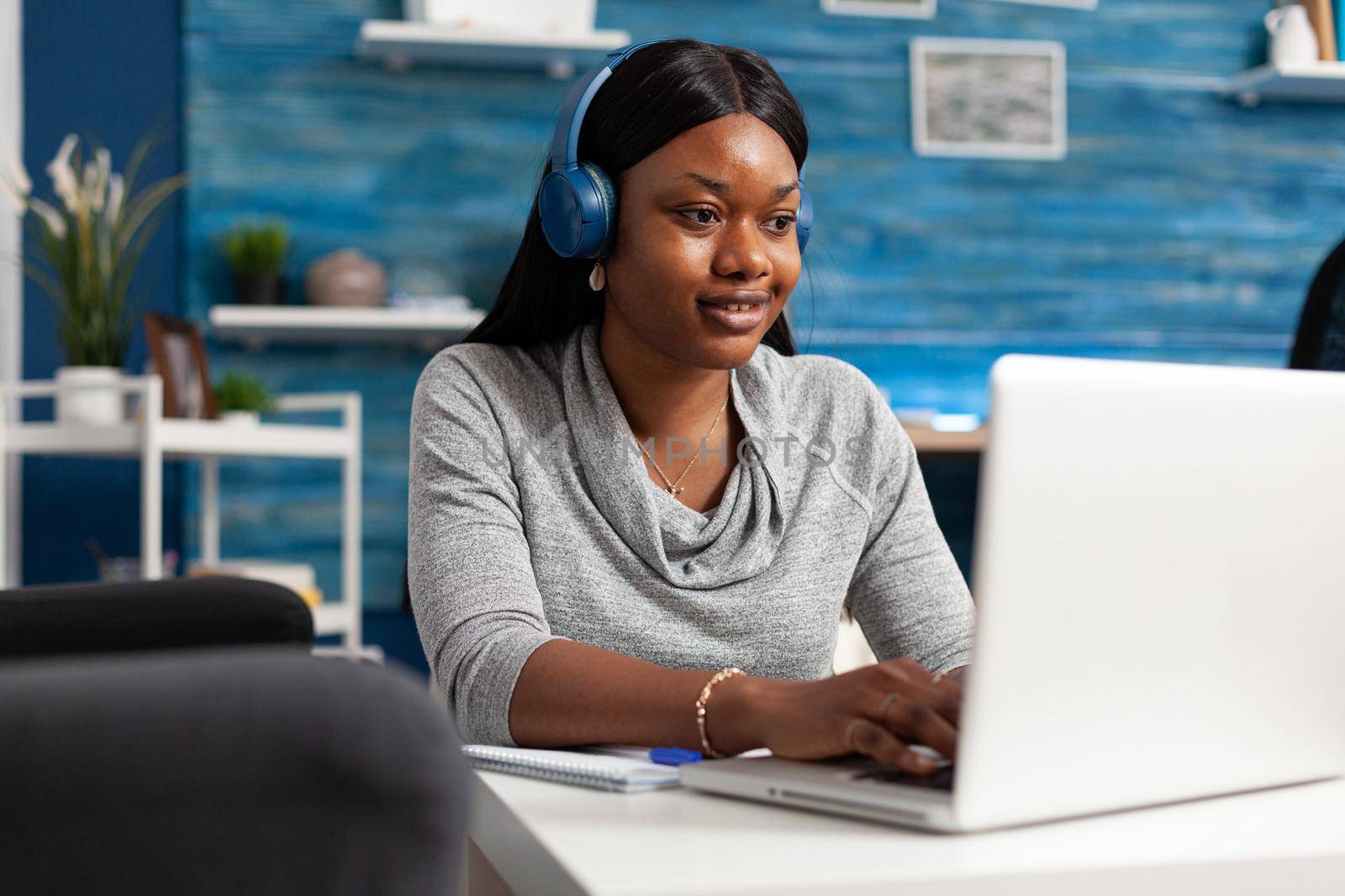 African american student wearing headphones browsing information composing blog article using laptop computer. Black woman typing education email working at social media webinar in living room