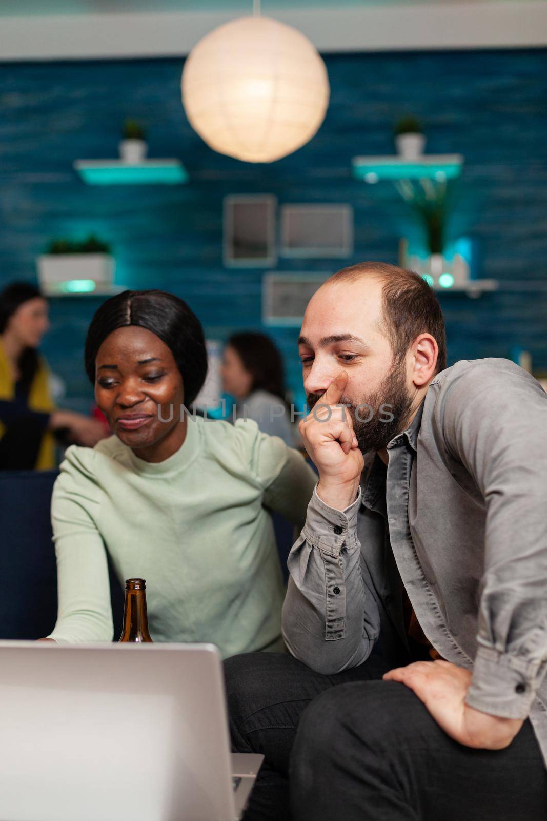 Multi ethnic friends socializing celebrating friendship watching funny video on laptop sitting on couch. In background two women drinking beer enjoying time spend together during entertainment party.