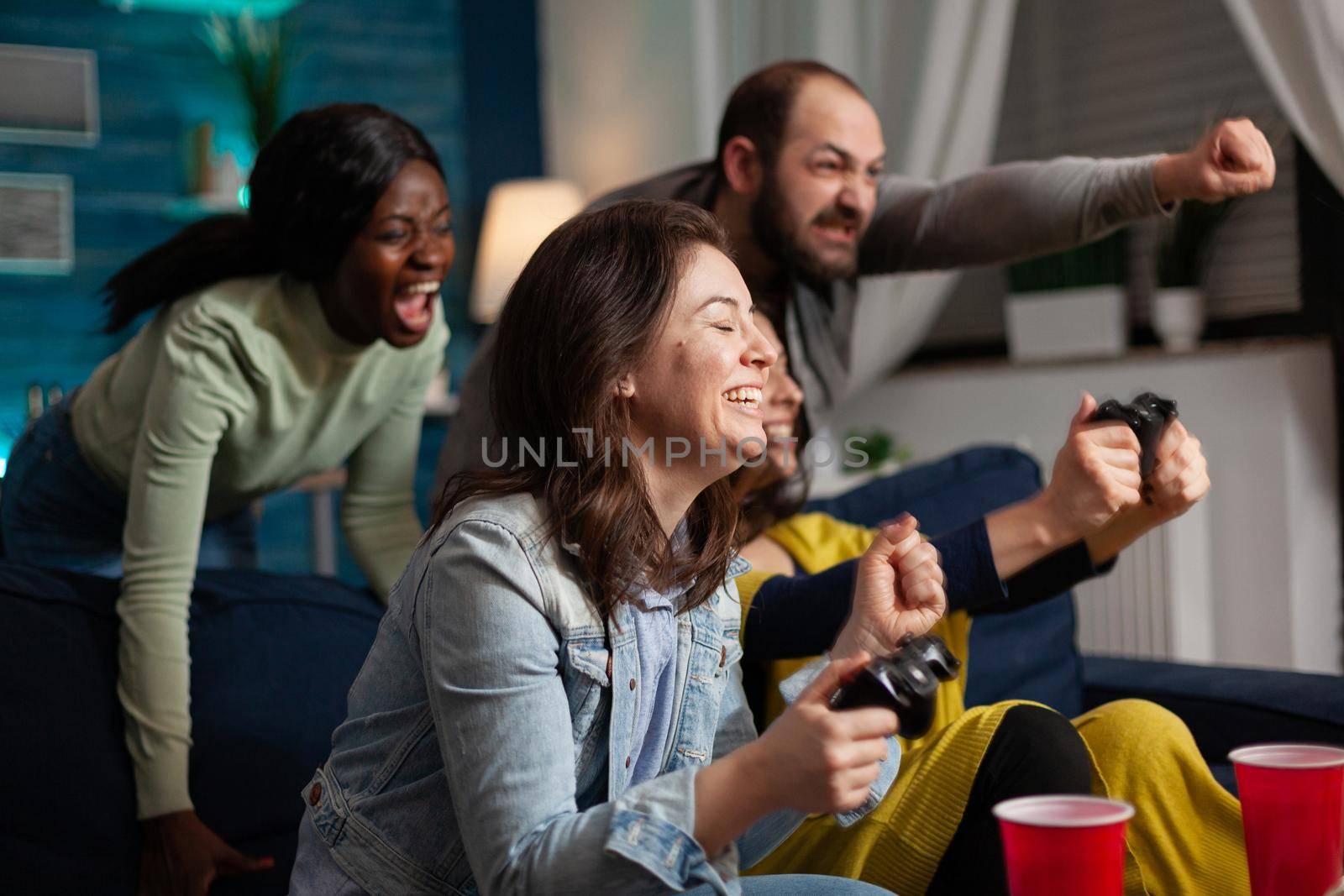 Excited group of multi ethnic friends celebraing victory aftter winning at online video games using wireless controller, late in the evening socialising sitting on couch.