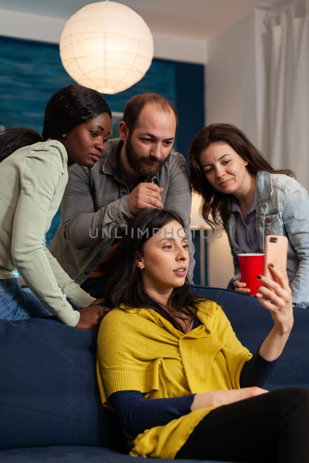 Multi ethnic friends having a virtual video call on smartphoneand bonding. Group of multiracial people spending time together sitting on couch late at night in living room.