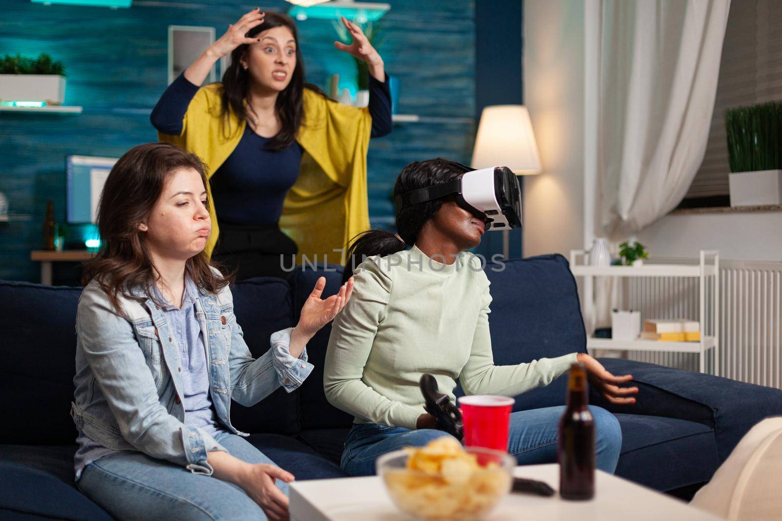 Upset multi ethnic women after losing while playing video games wearing virtual reality goggles. Mixed race group of people hanging out together having fun late at night in living room.