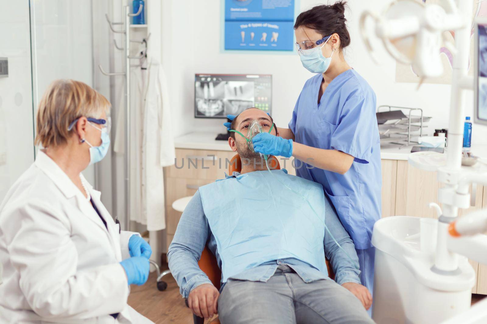 Sick man preparing for tooth surgery while nurse putting medical oxigen mask sitting on dental chair during stomatology hospital office. Dentist senior woman examining toothache treatment