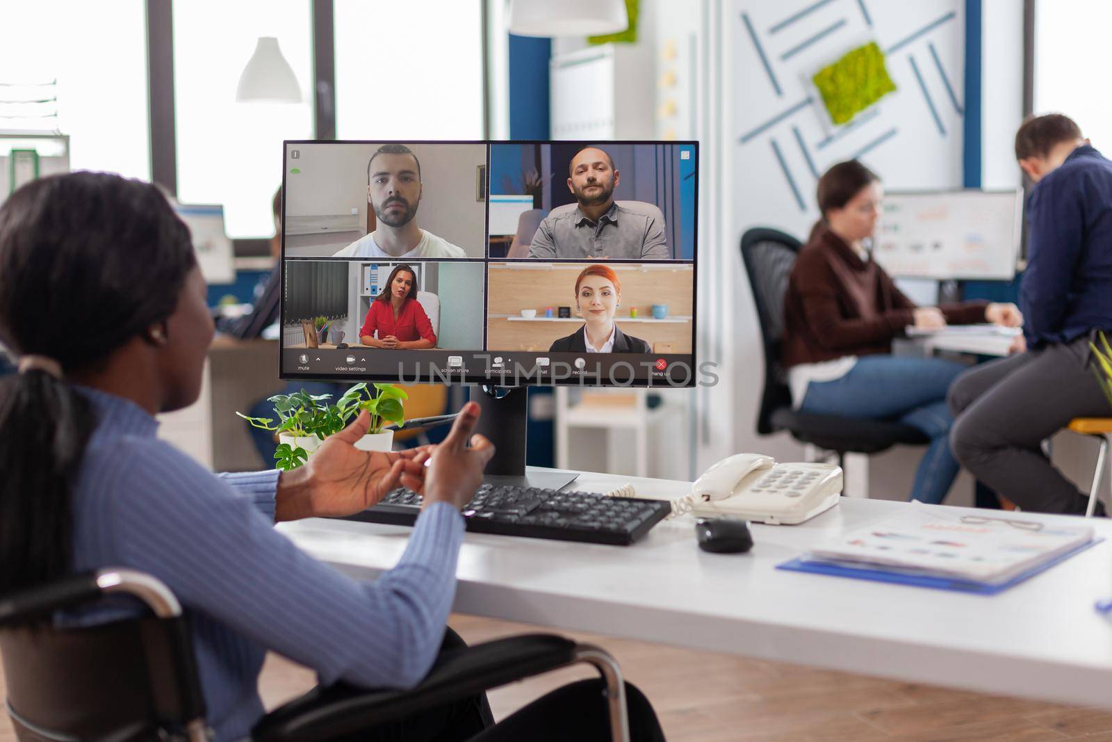 Paralysed handicapped black worker sitting immobilized in wheelchair having videomeeting discussing online with remotely colleagues in business office. Invalid employee working in financial company.