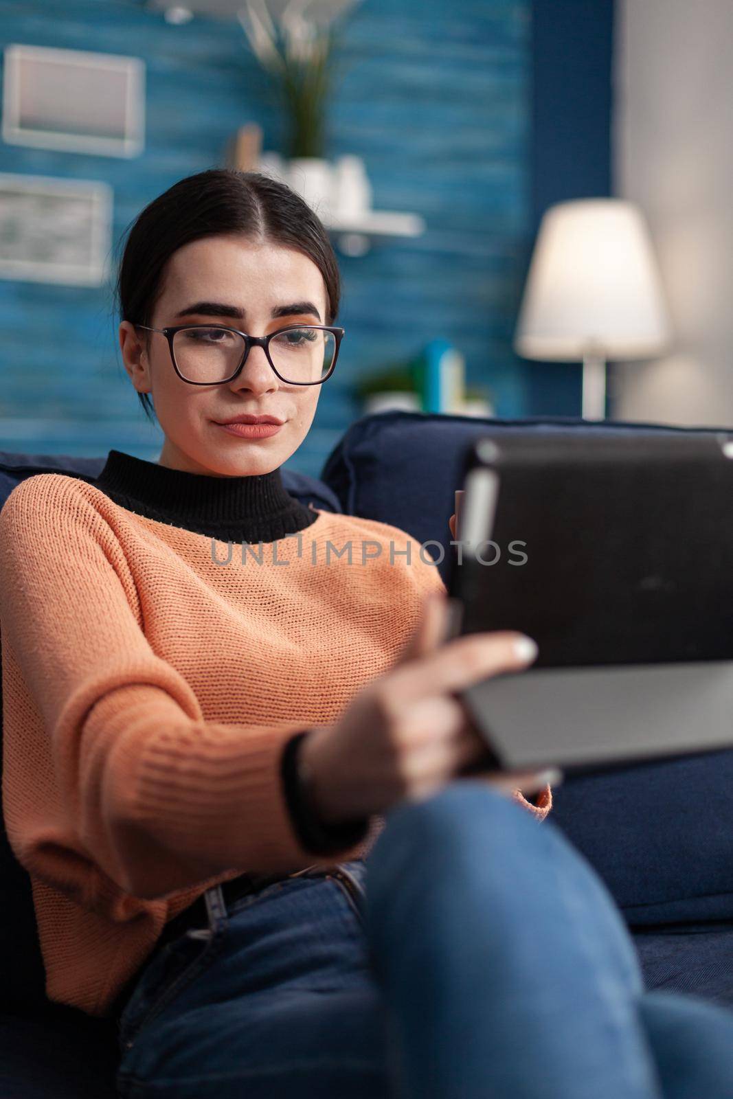 Portrait of student looking at tablet computer, sitting on sofa in living room drinking coffee. Teenager studying social communication using university digital app on mobile device