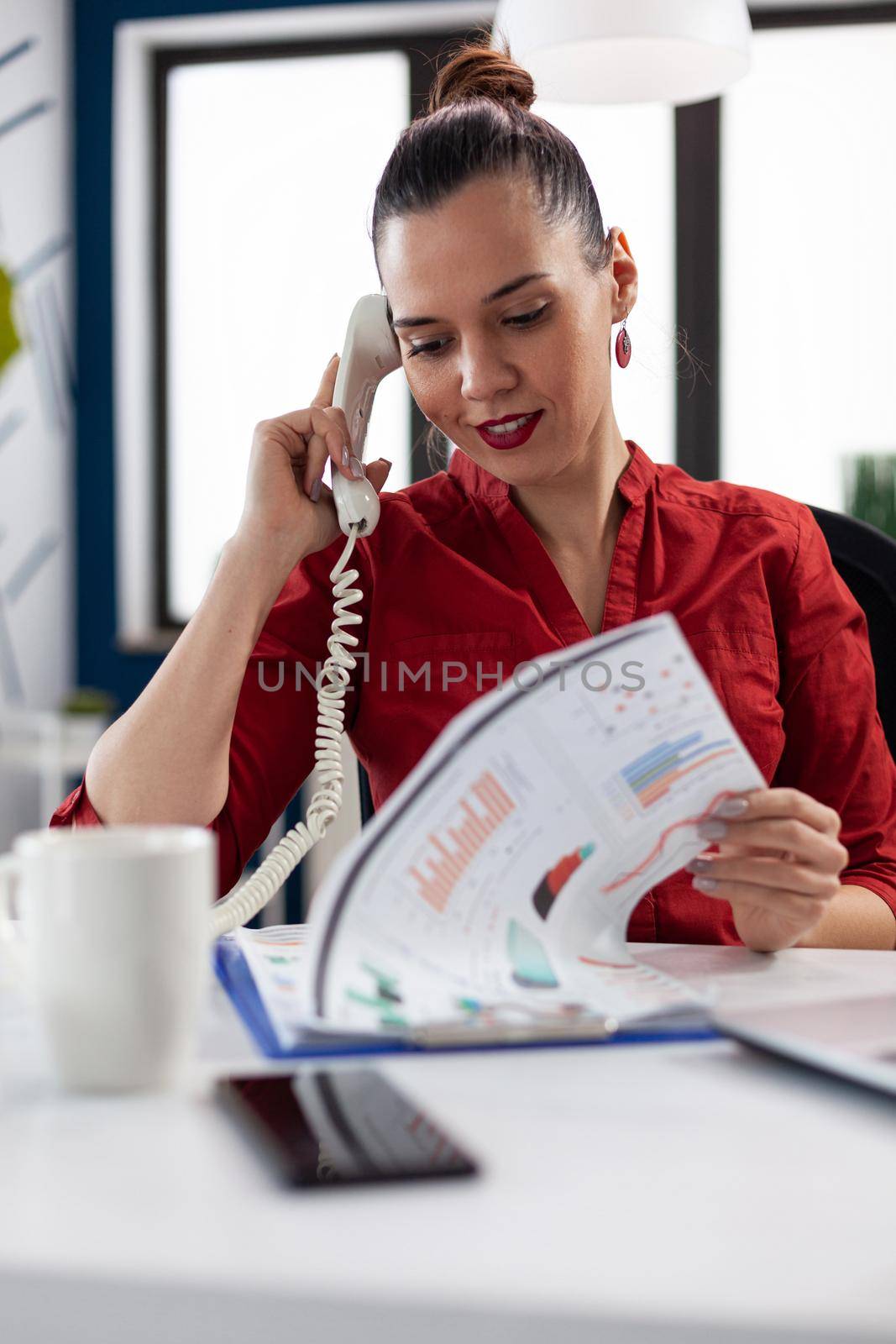 Businesswoman looking through finance charts on clipboard, making a call discussing with client sitting at desk in corporate workplace office. Manager giving finance expertise through phone call.