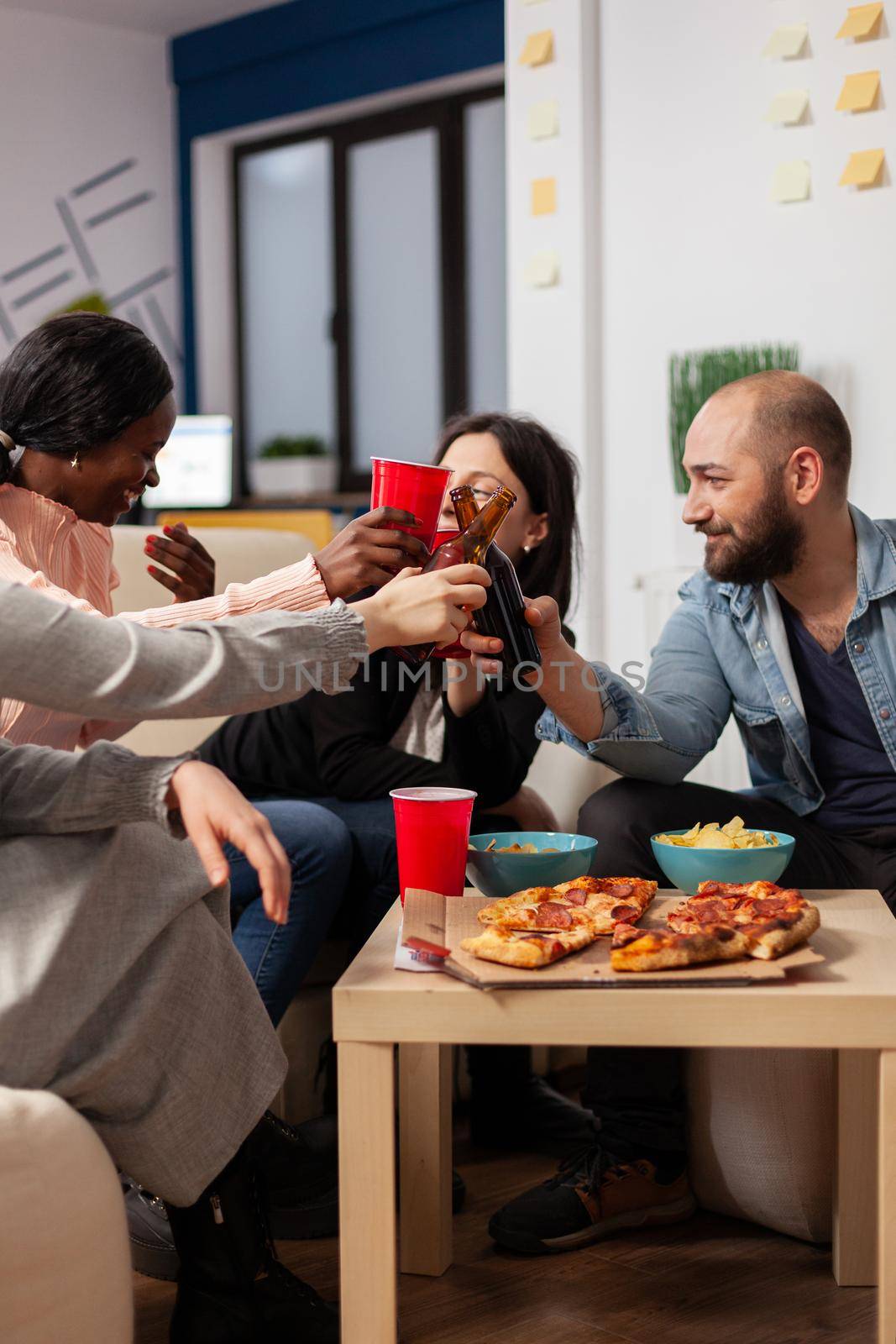 Diverse group of workers having fun after work at office meeting party. Cheerful friends cheers bottles and cups of beer to celebrate break from business. Multi ethnic people smiling