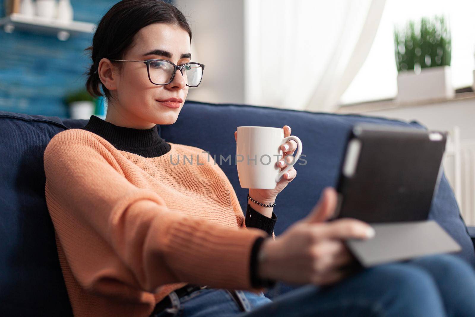 Student looking at online course about social media communication using mobile device computer, sitting on couch in living rom drinking cup of coffee. Teenager reading university communication online internet email