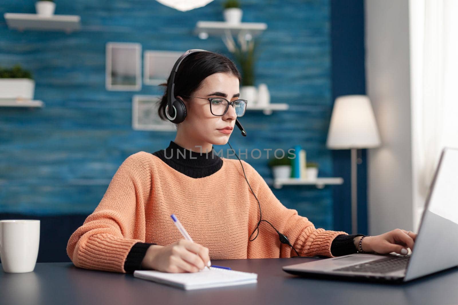 Student with headphones writing notes on notebook while searching communication information using laptop computer. Concentrated teenager making homework while sitting at desk in living room
