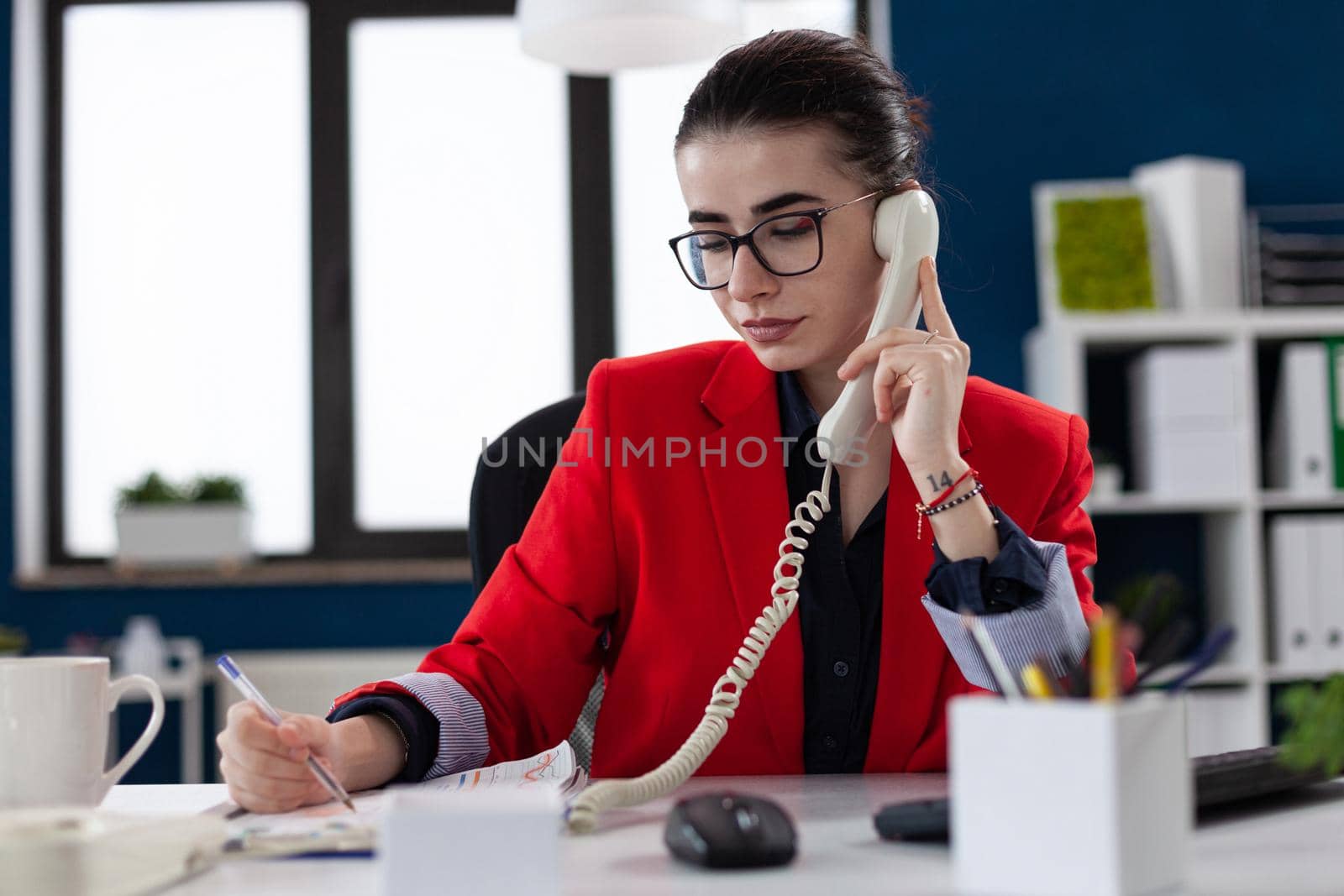 Businesswoman taking notes on clipboard sitting at desk in corporate office, while taking with adviser about financial expertise. Busy manager having a conversation about deadline.