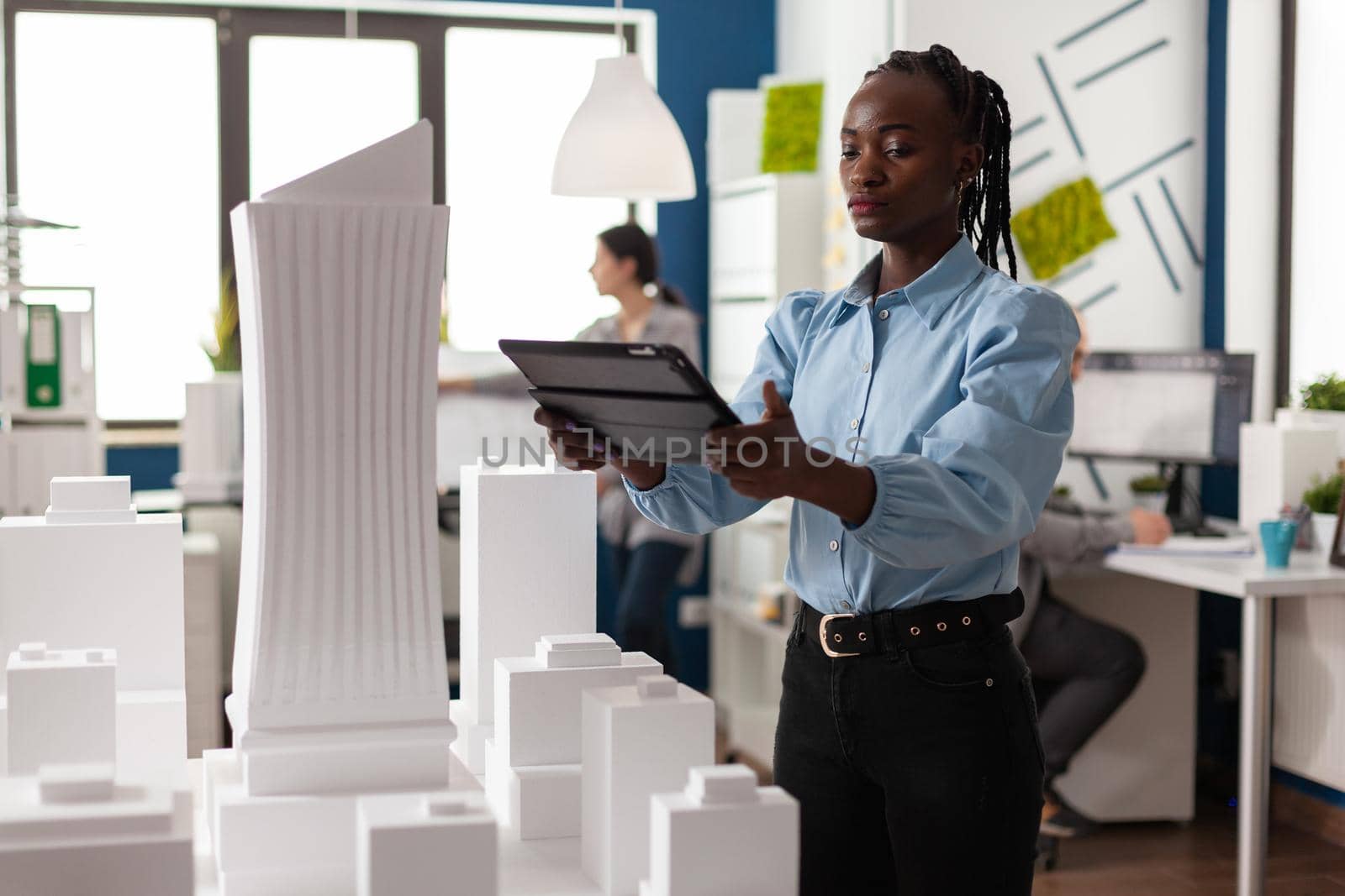 Architect woman of african american ethnicity working on tablet looking at professional maquette building model. Architectural worker viewing design for modern project in development