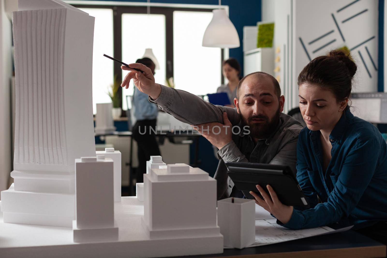 Team of construction architects work on tablet at office to plan building model maquette. Caucasian engineering partners pointing at layout project to design modern digital blueprint