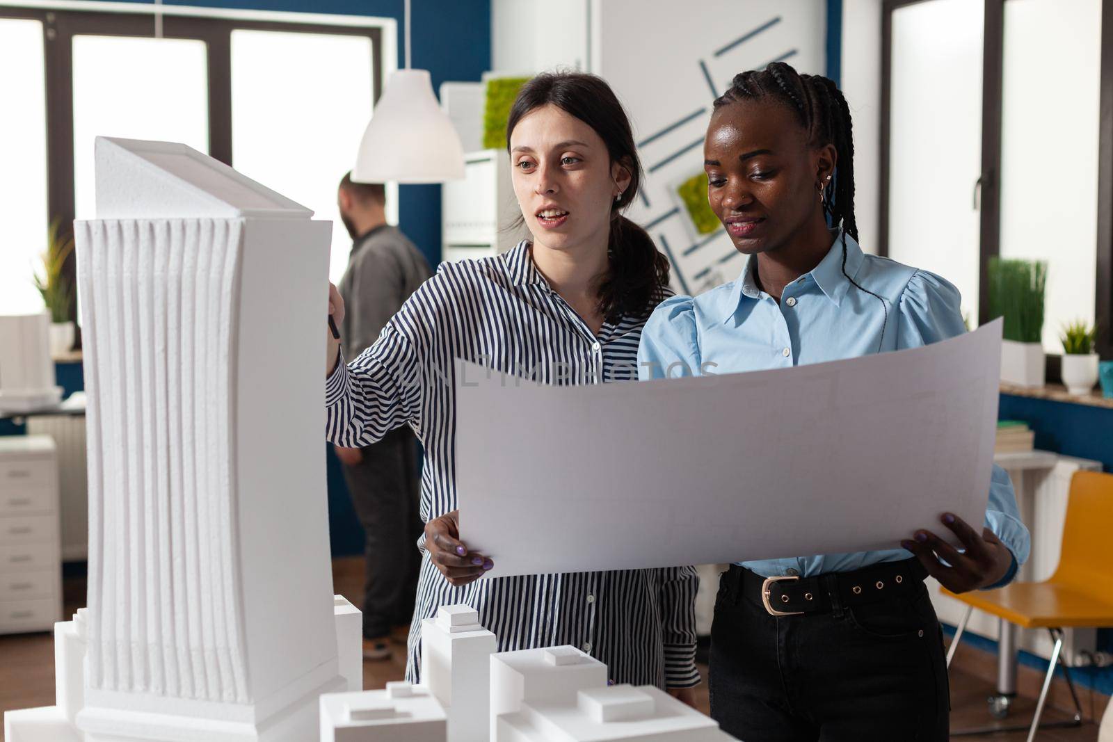 Colleagues architects women checking plans of blueprints layout design. Multi ethnic team of professional workers standing at building model maquette for modern technology project