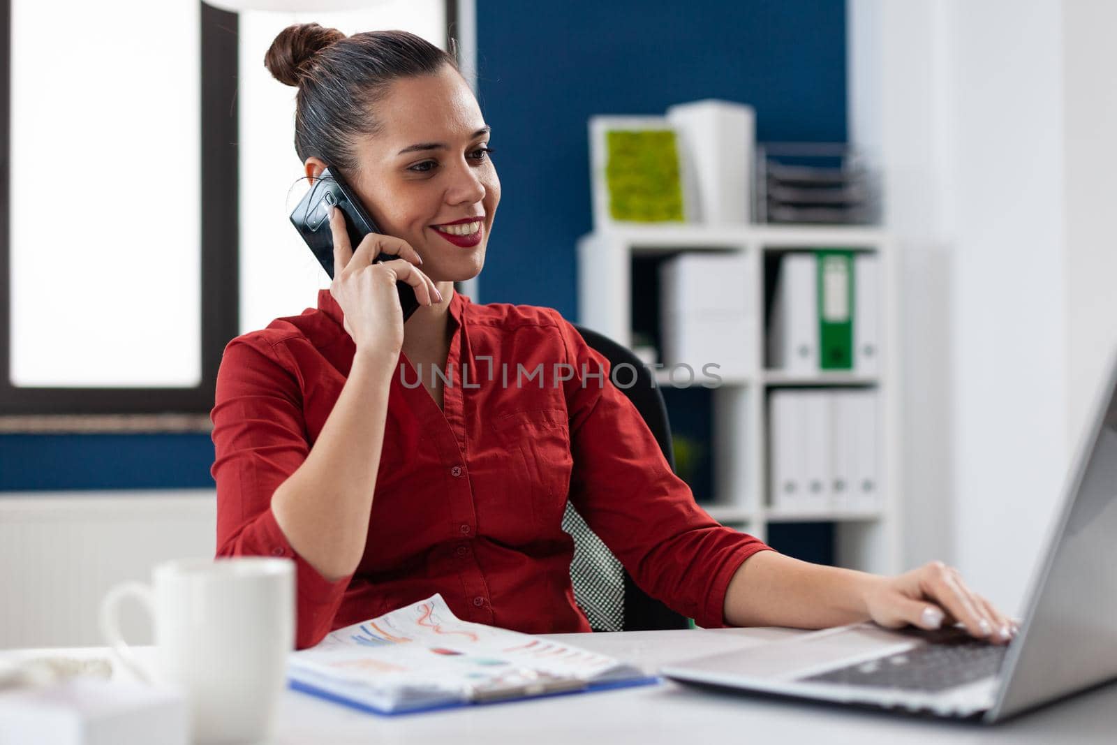 Busy businesswoman answering phone call sitting at desk in corporate workplace , discussing with client. Entrepreneur manager discussing with colleagues during call, looking at laptop screen.