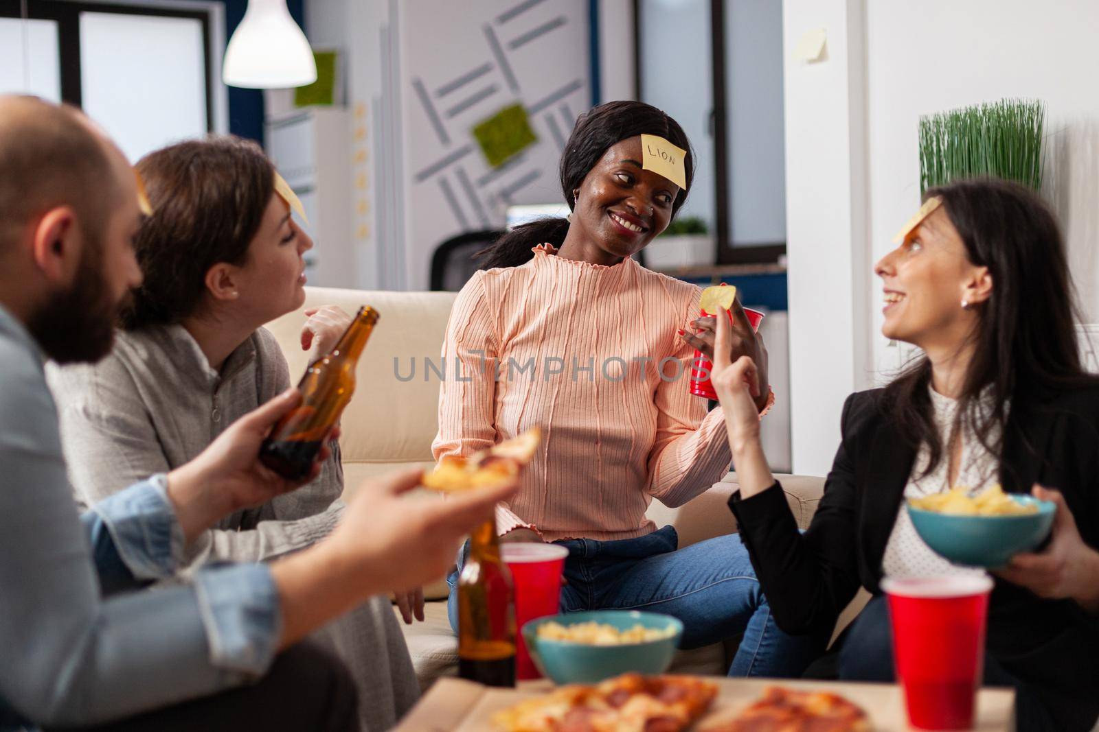 African american woman playing guessing game with friends after work at office. Multi ethnic group of workers guess imitation for fun cheerful activity while eating and drinking beer