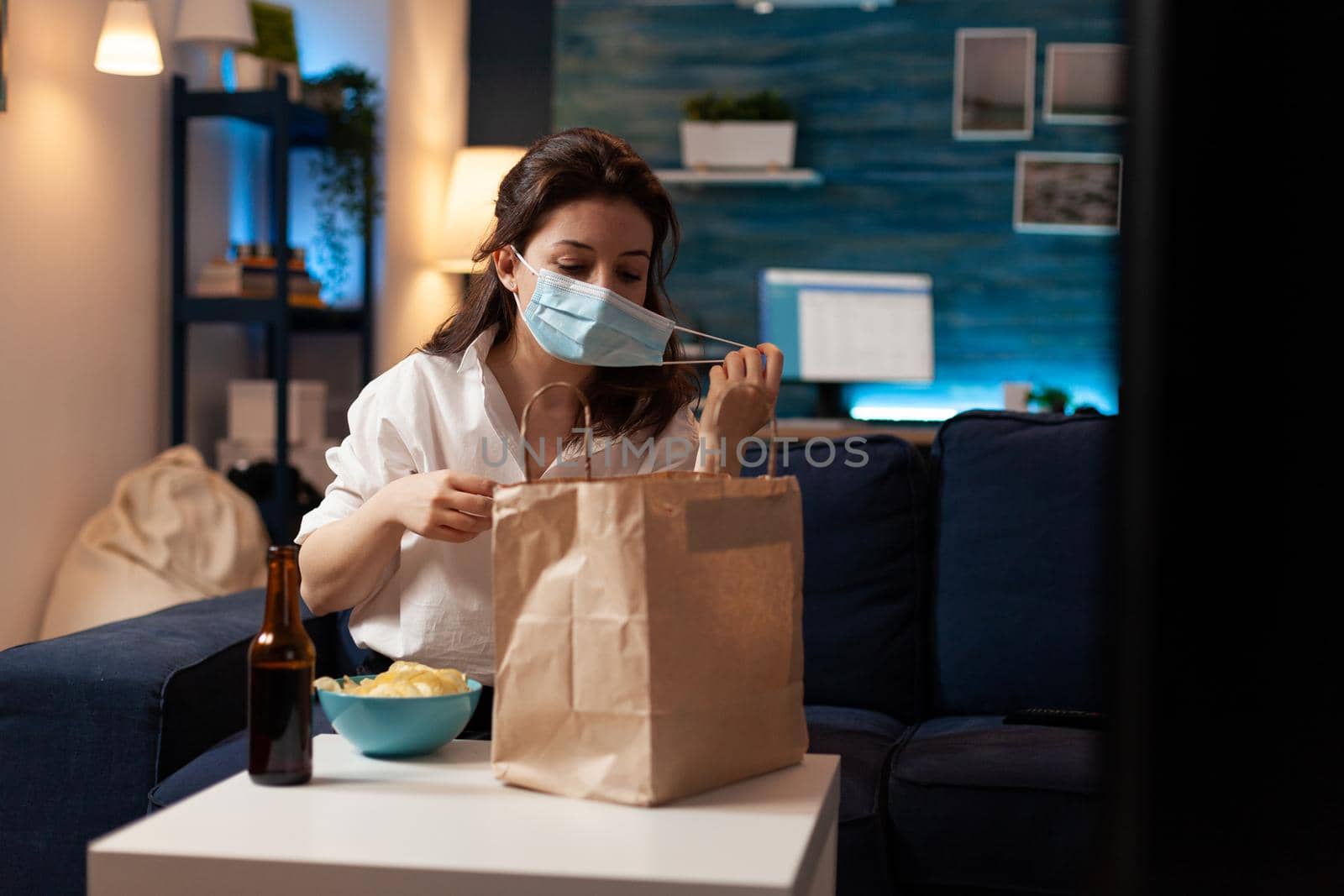 Cheerful woman taking medical protection face mask out after purchasing takeaway food in evening. Smiling caucasian female enjoying delicious junk-food home delivered. Tasting box order