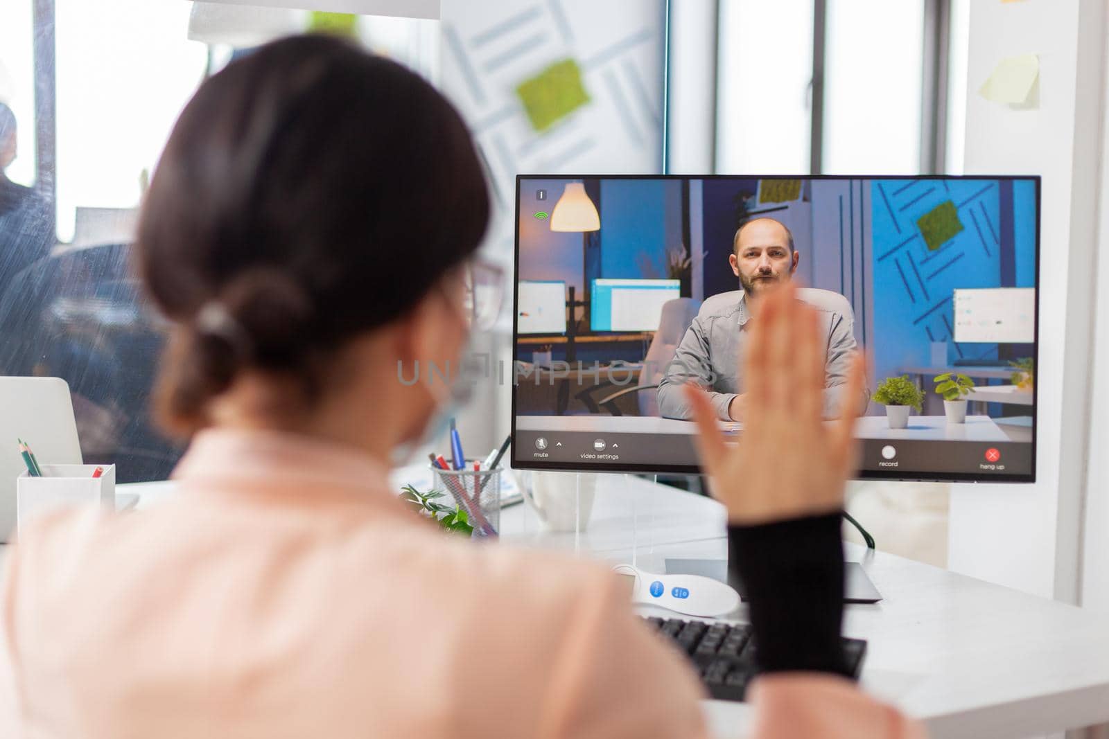 Woman greeting businessman during video call from new normal business ofice, in time of covid19 outbreak. Coworkers discussing project on remote call wearing face mask as safety prevention.