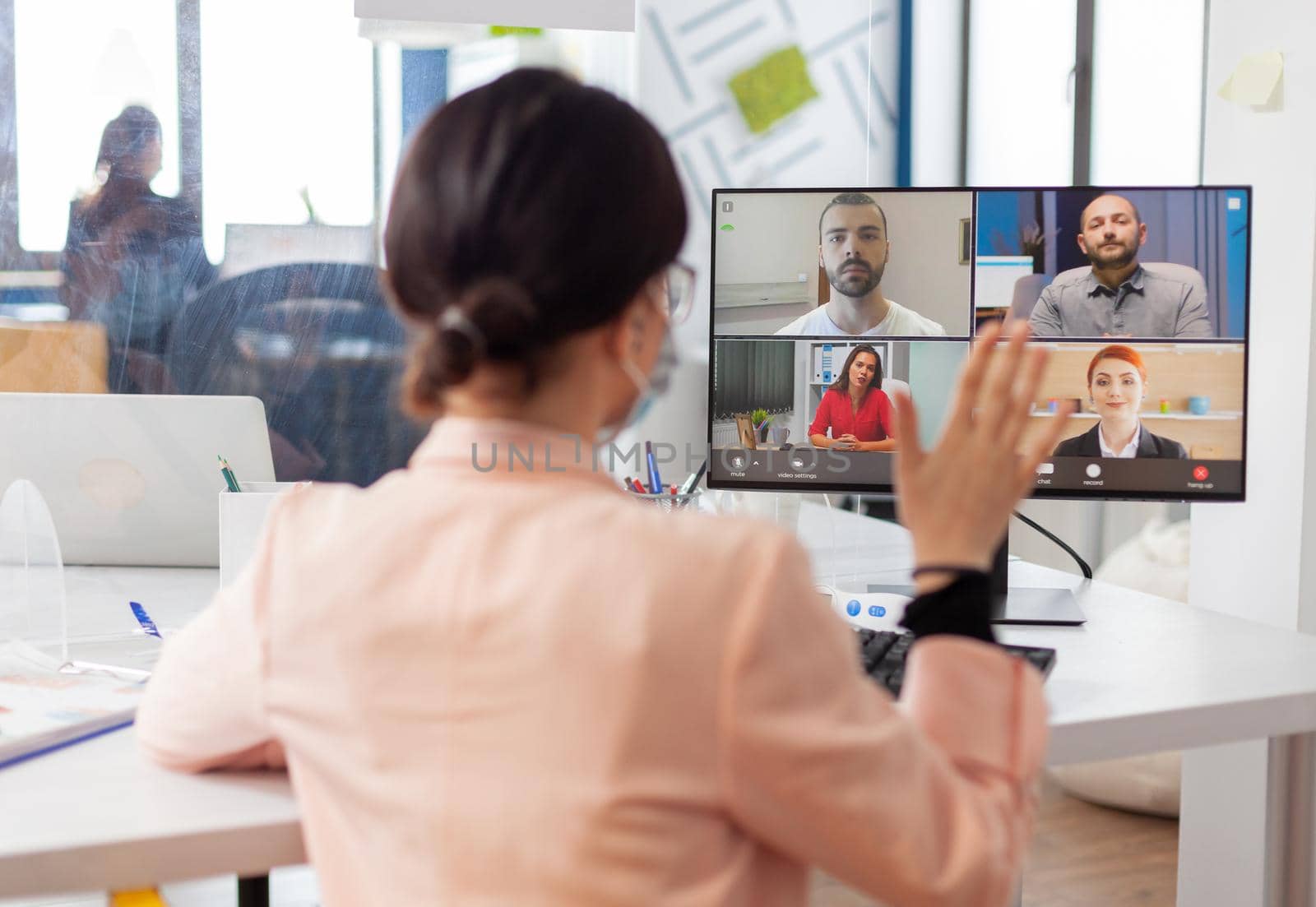 Executive woman in new nomal workplace talking on remote video call with team, wearing face mask due to coronavirus flu outbreak. Looking at monitor screen during virtual call.