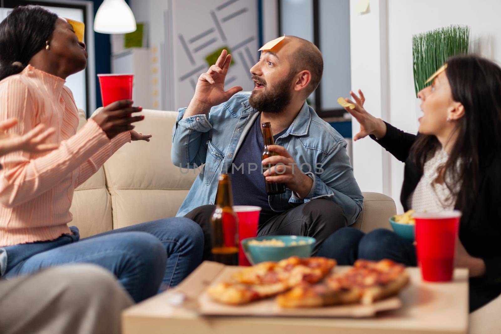 DIverse team of coworkers playing charades after work at office space. Multi ethnic friends enjoy guessing game of imiation as fun cheerful activity entertainment with food and drinks