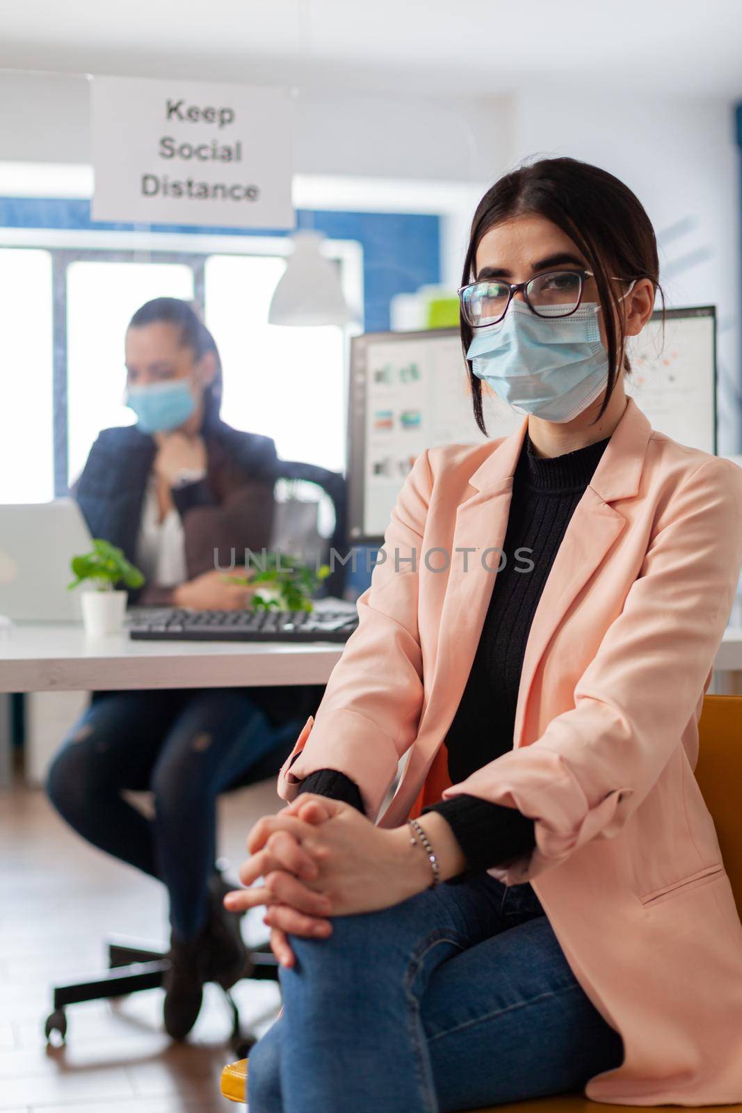 Portrait of manager entrepreneur with face mask as safety precation during coronavirus outbreak keeping social distancing from coworkers in office building looking at camera.