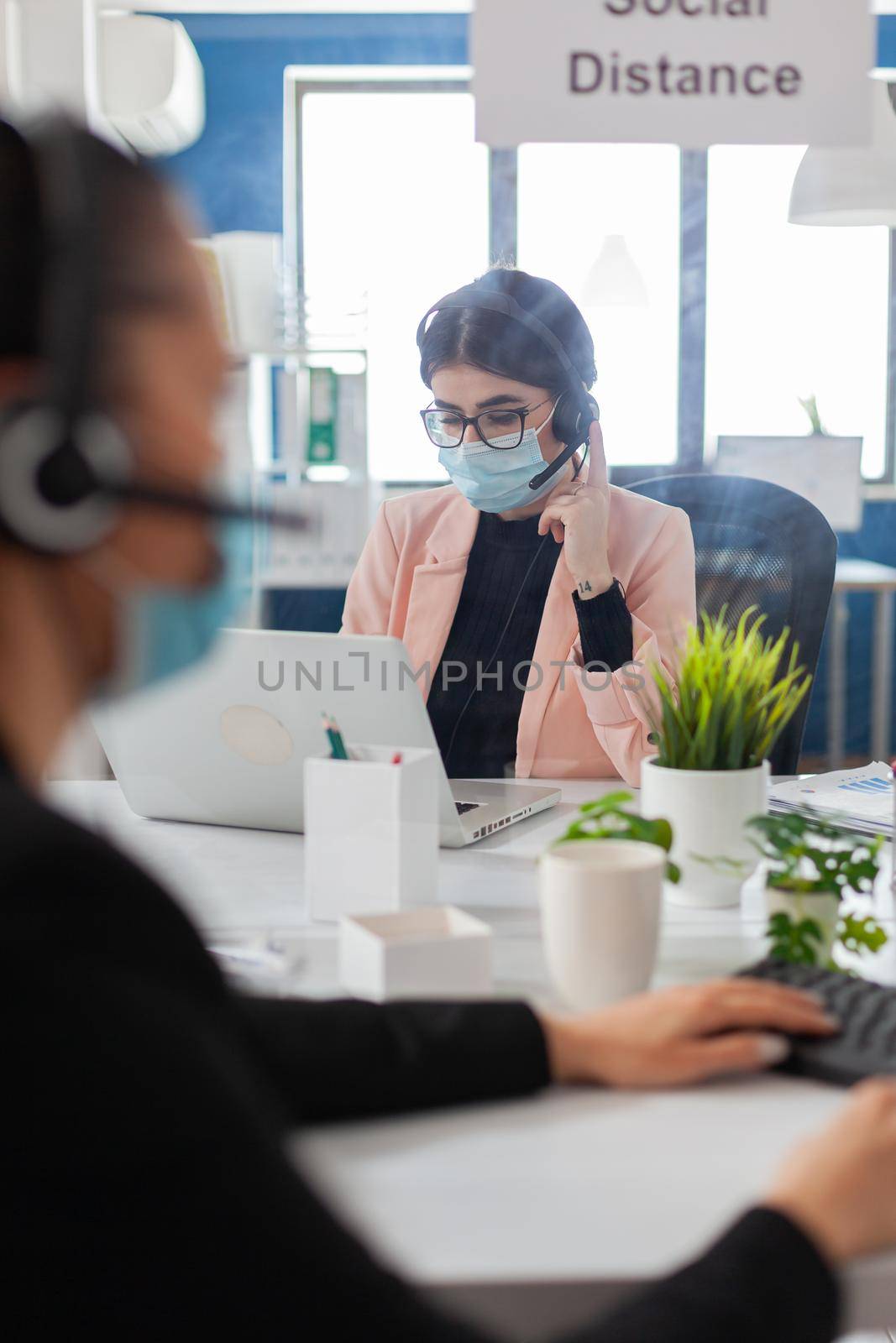 Business workers talking with clients using headphones in workplace, wearing face mask as prevention to coronavirus during global pandemic, keeping social distancing with shield.